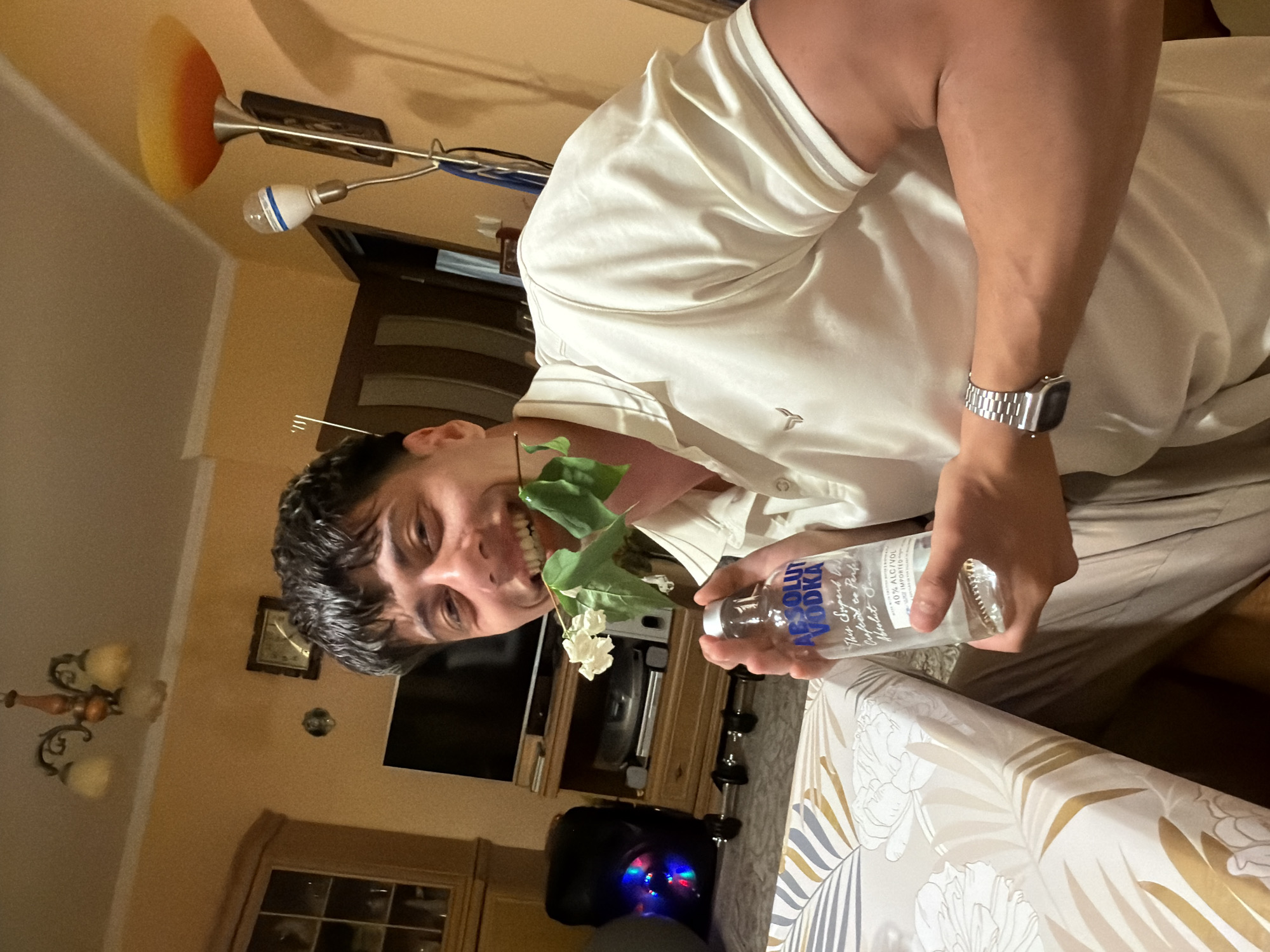 The image shows a man indoors, possibly at a residence, holding a flower in his mouth and a bottle of Absolute Vodka in his hand. The environment is indoors, with a table covered in a floral-patterned tablecloth in the foreground. In the background, there is a television set, furniture, and lighting fixtures, indicative of a home setting. The man has dark hair and is wearing a watch on his left wrist. The bottle is labeled with the words "ABSOLUT VODKA" and "IMPORTED." There is no direct evidence in the image to indicate the location is Burgas, Bulgaria.