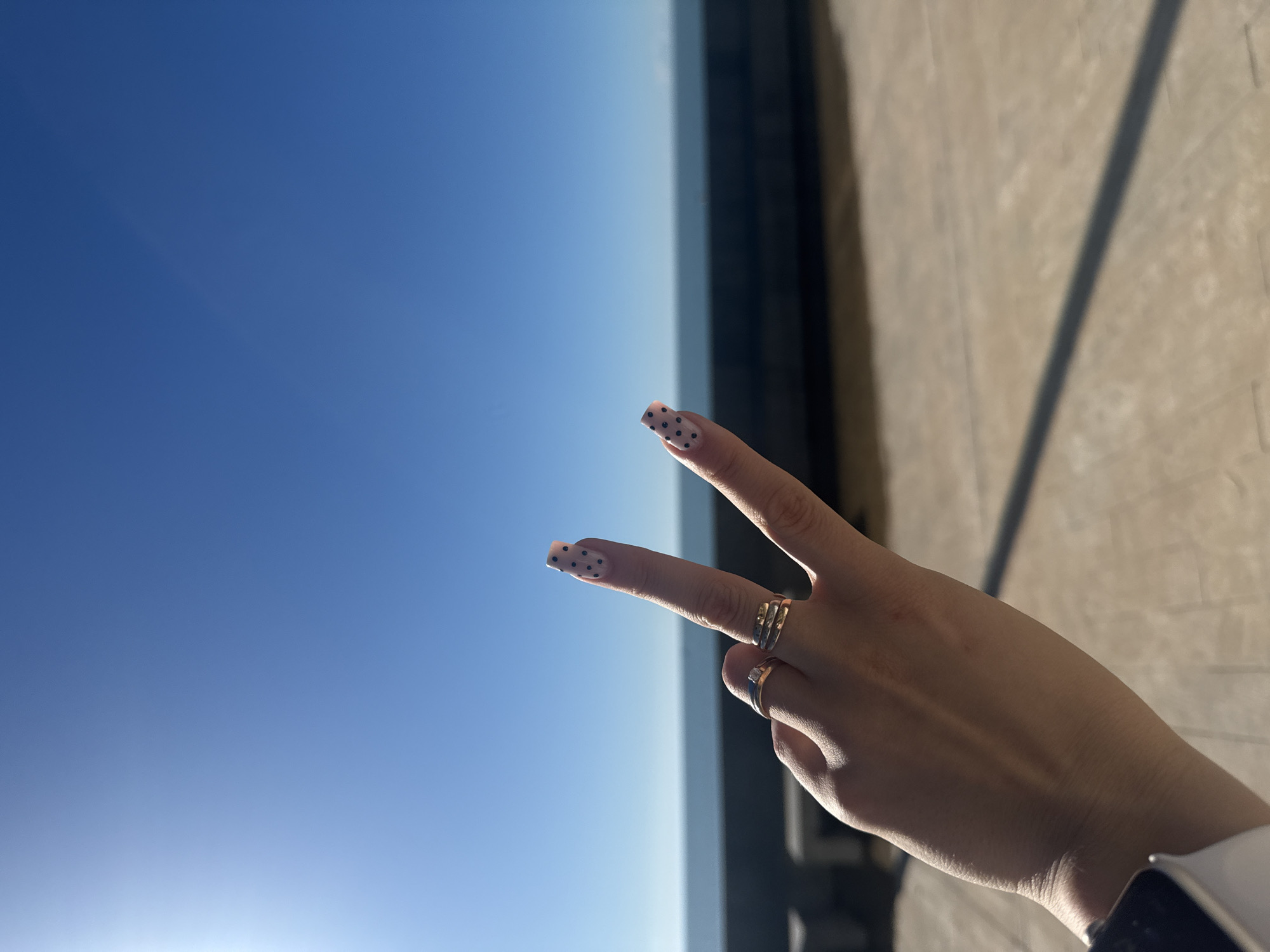 A person's hand is raised in a peace sign against a backdrop of a clear blue sky and the edge of what appears to be a building. The hand has two rings on the ring finger and middle finger. The fingernails are painted with a light color and have black polka dots. The background consists of a gradient blue sky that transitions from light to dark, and on the right side, there is the edge of a beige colored wall in the background. The building wall is cast in shadow. The location is Batumi, Georgia.