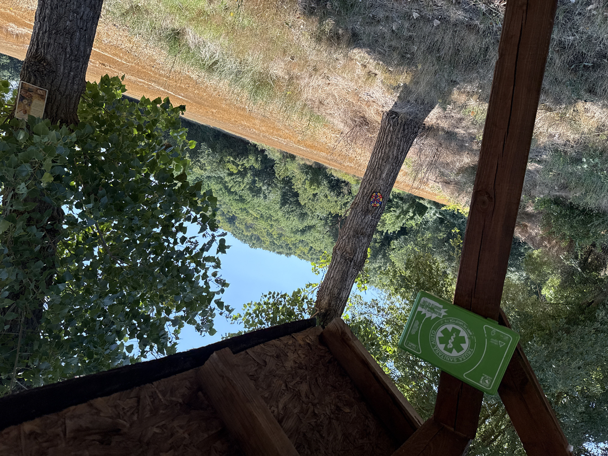 The image depicts an outdoor scene in Pravdino, Bulgaria, under a clear blue sky. The foreground features a wooden structure, possibly part of a shelter or an informational kiosk, with a green first-aid kit attached to a wooden support beam. This kit bears Italian writing and a medical symbol. To the left of the structure, a large tree trunk is visible, also a wooden post.
In the midground, a field of dry, golden grass or crop stretches diagonally across the scene. Two trees stand within this field. The tree on the left has a colorful circular emblem attached to its trunk. The tree on the right has a small framed picture or religious icon affixed to its bark.
The background consists of a densely wooded hillside, suggesting a rural or natural setting. The overall impression is one of a remote or nature-oriented location, potentially a hiking trail or a rest stop. No people are present in the image. The lighting indicates it is daytime, with shadows cast by the wooden structure and trees, implying the sun is positioned to the right.