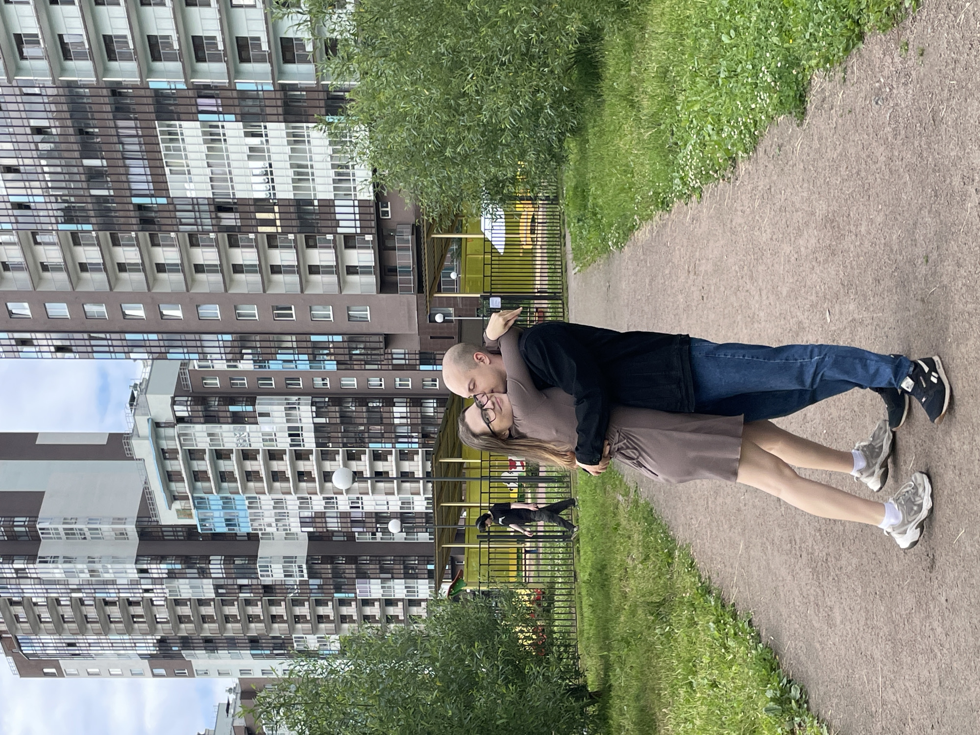 Two people, a man and a woman, are embracing on a paved path in a residential area. The man is wearing a black t-shirt and blue jeans, while the woman is wearing a light brown dress. Both are wearing athletic shoes and white socks. The man has his arm around the woman, and they are leaning into each other. The woman is wearing glasses and has her eyes closed. They appear to be in a romantic moment.
Behind them, to the left, is a modern, multi-story apartment building with many windows. To the right of the path is a grassy area with some small white flowers and a row of trees. Further in the background, there is a fence and a playground structure with a yellow roof and some green elements.
The scene appears to be set during the daytime, under an overcast sky. The overall atmosphere is casual and relaxed, suggesting a moment of connection between the couple. The presence of the apartment buildings, playground, and grassy areas indicates an urban residential setting, consistent with the provided location context of Saint Petersburg, Russia. There is no visible text in the image.