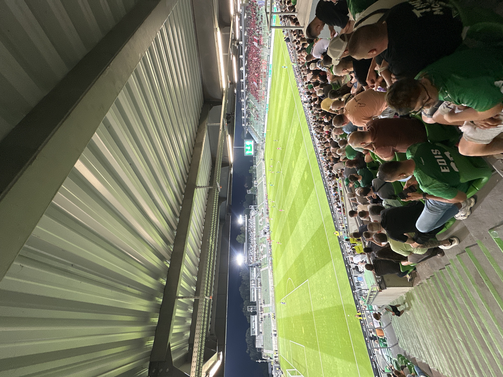 The image shows a section of a football stadium with a crowd of spectators seated on green bleachers. The field, illuminated by floodlights, is visible in the background, with a game in progress. Spectators are wearing a variety of casual clothing, with many in green attire. The stadium features a covered seating area with a corrugated metal roof. The atmosphere appears to be that of a typical football match with people engaged in watching the game. This scene is located at the Huvepharma Arena in Razgrad, Bulgaria, the home ground of PFC Ludogorets Razgrad.