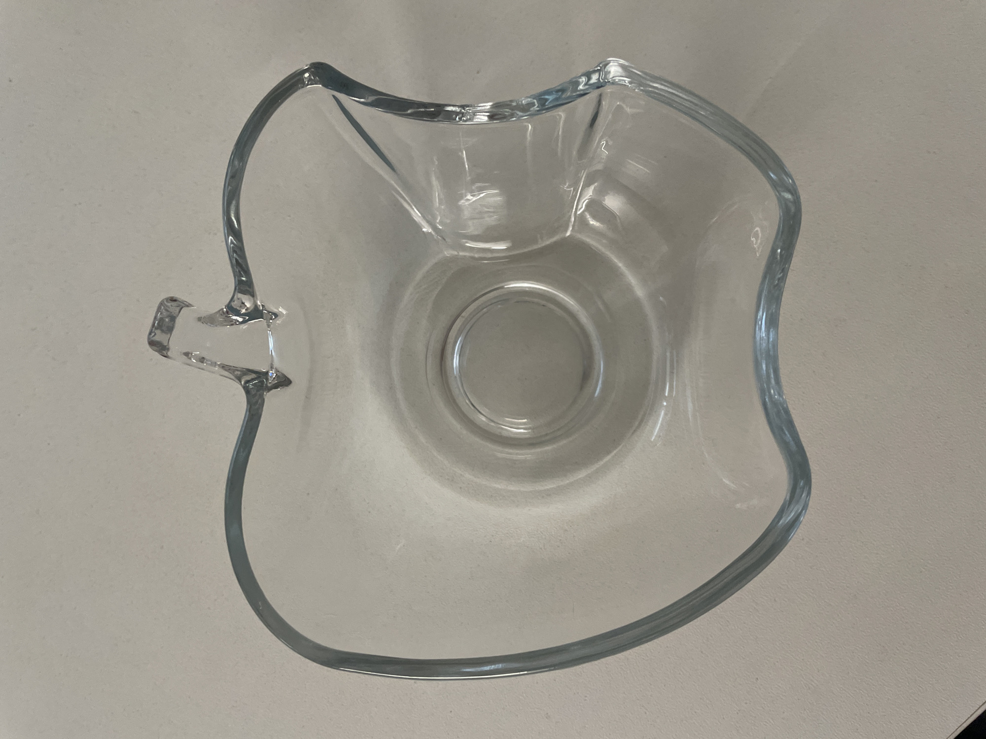 A clear glass bowl shaped like an apple sits on a light-colored, speckled surface, possibly a countertop. The bowl has a stem-like protrusion at the top and a divot on the right side mimicking an apple's indentation. The surface it rests on is smooth and appears to be indoors. There are no people or animals present in the image, and no specific details about the location within Vologda, Russia, are discernible from the provided image. The lighting suggests it's daytime, with soft shadows cast by the bowl.