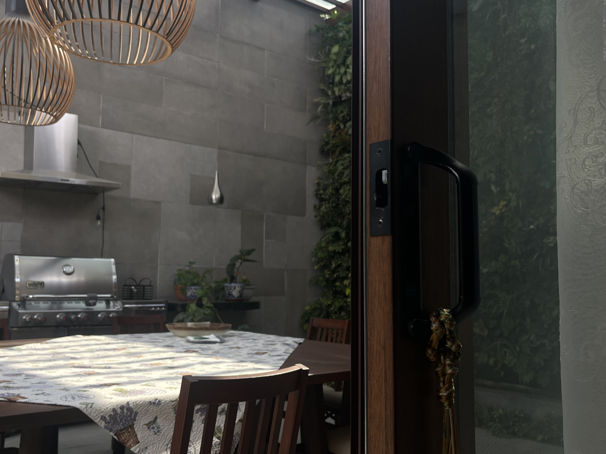 The image shows an outdoor dining area with a table and chairs, partially visible through a doorway. The table is covered with a tablecloth, and a grill is visible in the background. Behind the grill is a gray tile wall with plants growing on it. The doorway has a black handle. The location is San Bernardino Tlaxcalancingo, Mexico.