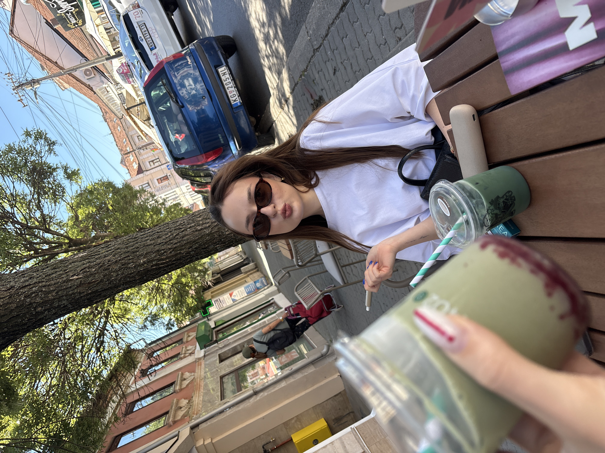 The image depicts a street-level view of a cafe in Kragujevac, Serbia, on a sunny day. A young woman with long brown hair and sunglasses is seated at a wooden table, pouting her lips towards the camera. She is wearing a white oversized t-shirt and holding a cigarette in her right hand. On the table are two green beverages in plastic cups with straws, one of which is being held by another person's hand, visible in the foreground. The hand has manicured nails with a white and red design. A black handbag rests on the table next to the woman. In the background, a blue Fiat Punto car is parked on the street, with its license plate reading "KG 205-NL". Further down the street, other cars and buildings with red-tiled roofs are visible. A person carrying a red wheeled bag and a backpack walks on the sidewalk. The overall scene suggests a casual outdoor cafe setting in an urban environment.