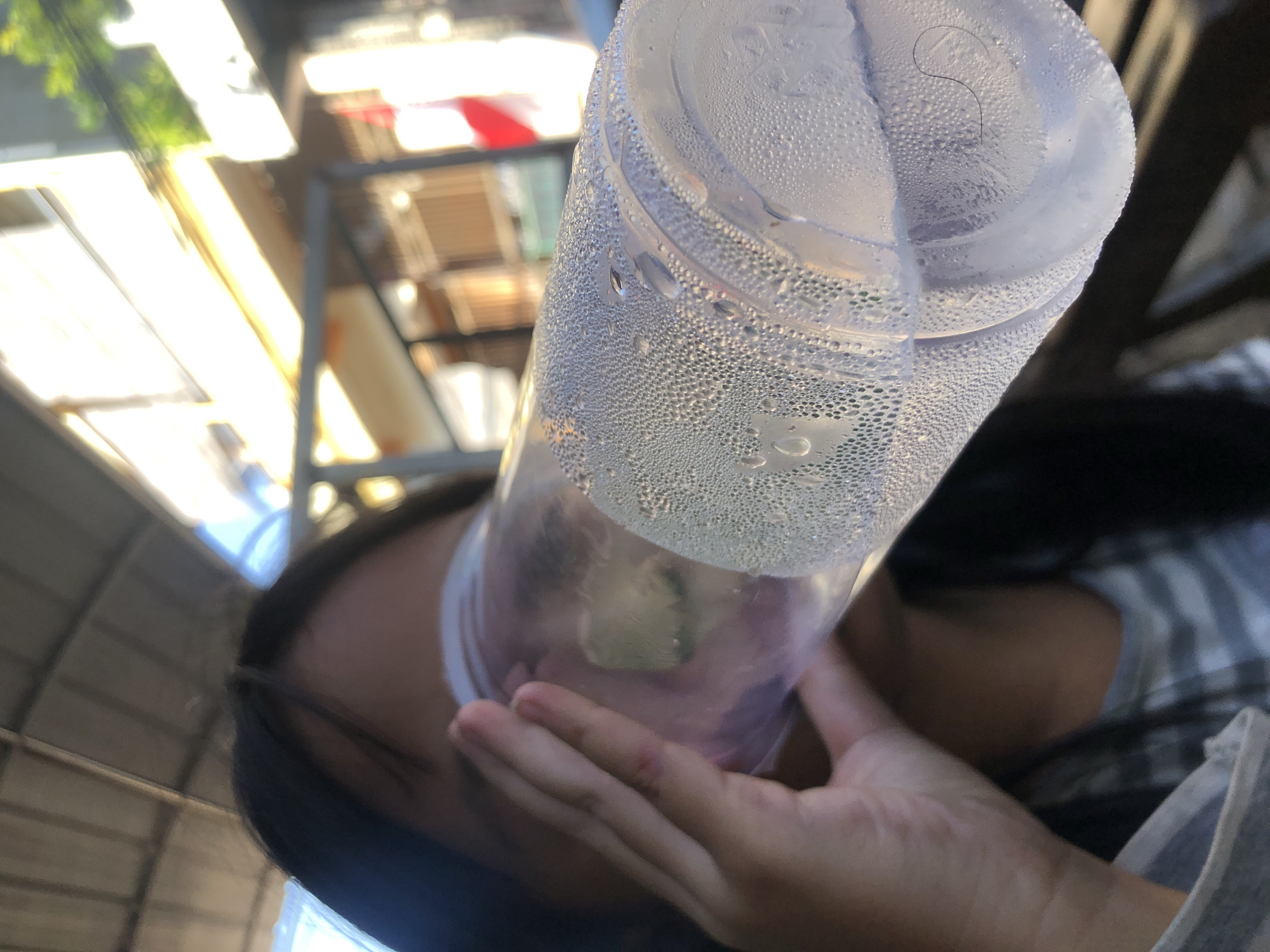A close-up, low-angle shot captures a person's hand and a portion of their face as they drink from a clear plastic cup filled with a light-colored liquid. The cup is covered in condensation. The background is blurred but suggests an outdoor setting with a canopy and structures that could be part of a building or market stall. The lighting indicates it is daytime, possibly late afternoon, with sunlight casting warm tones. While the image itself does not provide direct indicators of the location being Surabaya, Indonesia, the presence of a red and white flag, possibly the Indonesian flag, in the blurred background offers a potential clue. The overall impression is one of refreshment on a warm day.