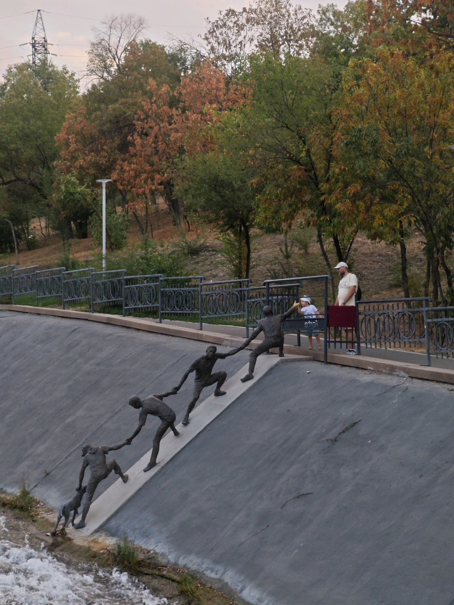 The image shows a public artwork depicting a group of people and a dog helping each other ascend a steep, gray embankment next to a body of water. The sculptures are made of dark metal and are arranged in a line, with each figure assisting the one behind them. A man and a child are standing on a walkway above the embankment, observing the sculptures. The surrounding environment features trees with autumn foliage, a metal fence, and a power line tower in the background. The scene is located in the city of Almaty, Kazakhstan, specifically in a park area along a waterway, possibly near a public promenade.