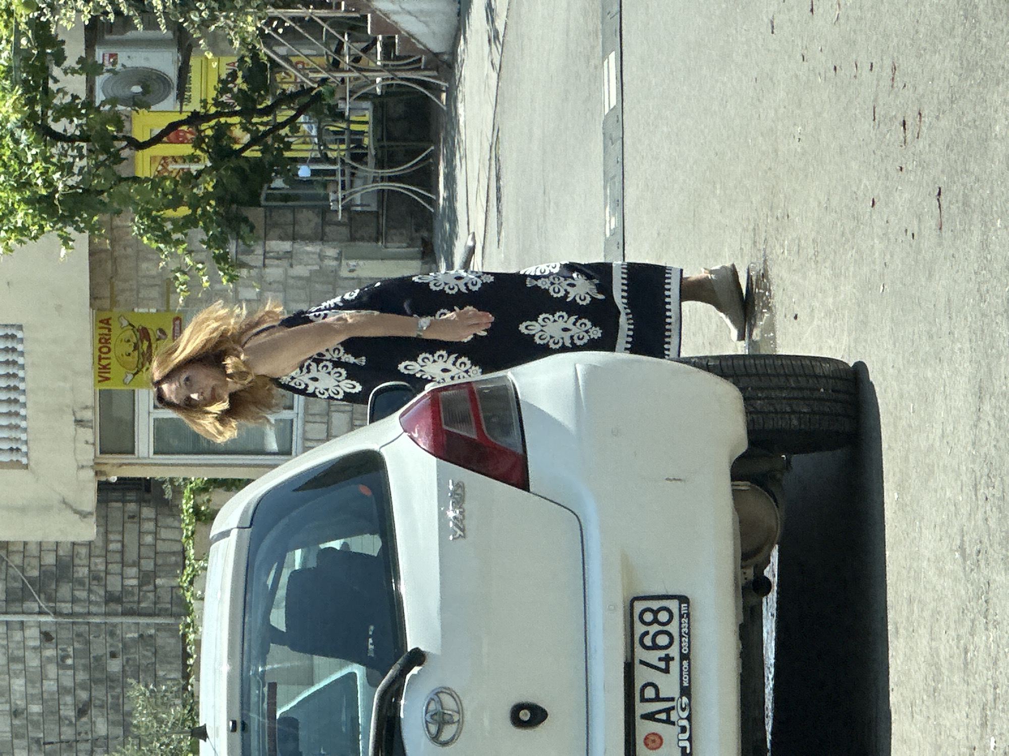 A white Toyota Yaris is parked on a street in Budva, Montenegro. The car is facing away from the camera, with its rear bumper and taillights visible. A license plate reading "AP 468" is attached to the bumper. A woman wearing a black and white patterned dress is standing next to the car, looking over her shoulder at the camera. She has long, blonde hair and is wearing a watch on her left wrist. The background shows a brick building with a yellow sign that says "VIKTORIJA." There are trees and outdoor seating areas visible in the background. The street is paved with asphalt, and there are some puddles on the ground.