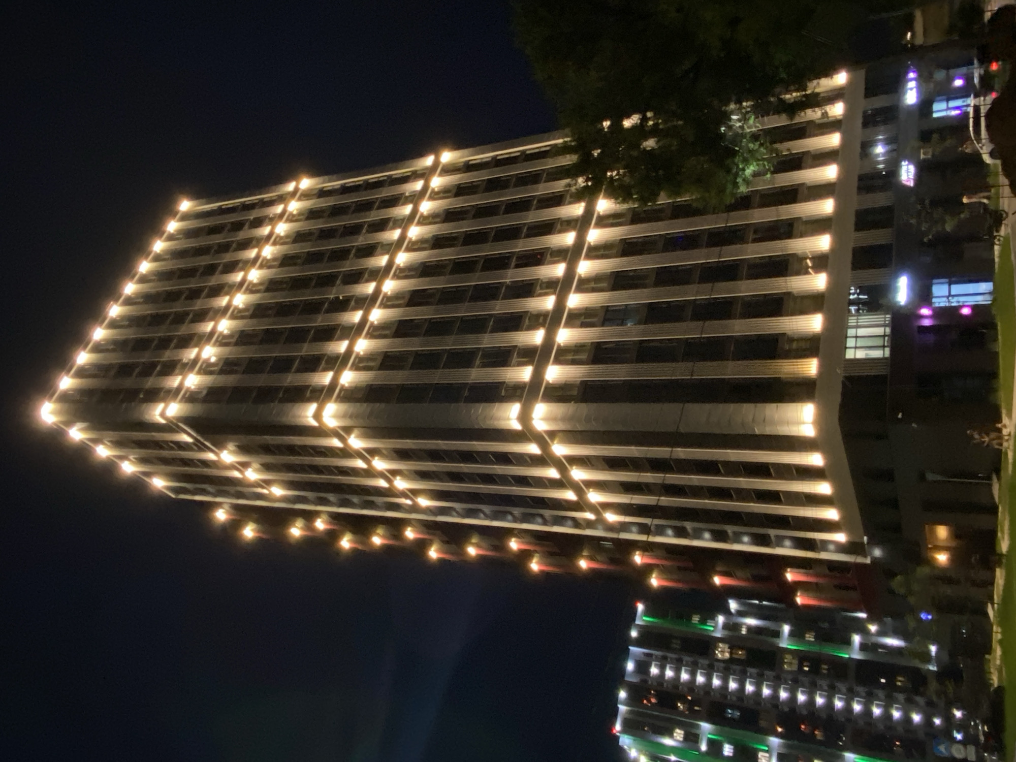 The image shows a modern, multi-story building illuminated with warm yellow lights at night. The building has a tall, rectangular structure with a facade characterized by vertical columns. Lights are strategically placed along the top edges and at the base of these columns, creating a dramatic, glowing effect against the dark night sky.
To the left, another building is visible, also illuminated, but with green and red lights, suggesting it may be a residential complex or commercial property with a different lighting scheme. Trees are present in the foreground, partially obscuring the lower levels of the main building. The ground level of the main building features storefronts with illuminated signage.
There are a few individuals visible on the sidewalk and near the entrance of the building, appearing to be pedestrians. Vehicles, including a white car, are parked or moving in the vicinity. The overall scene depicts an urban environment at night, highlighting the architecture and lighting of the buildings.
The image is attributed to Blagoveshchensk, Russia. The specific location is likely a modern development area within the city.