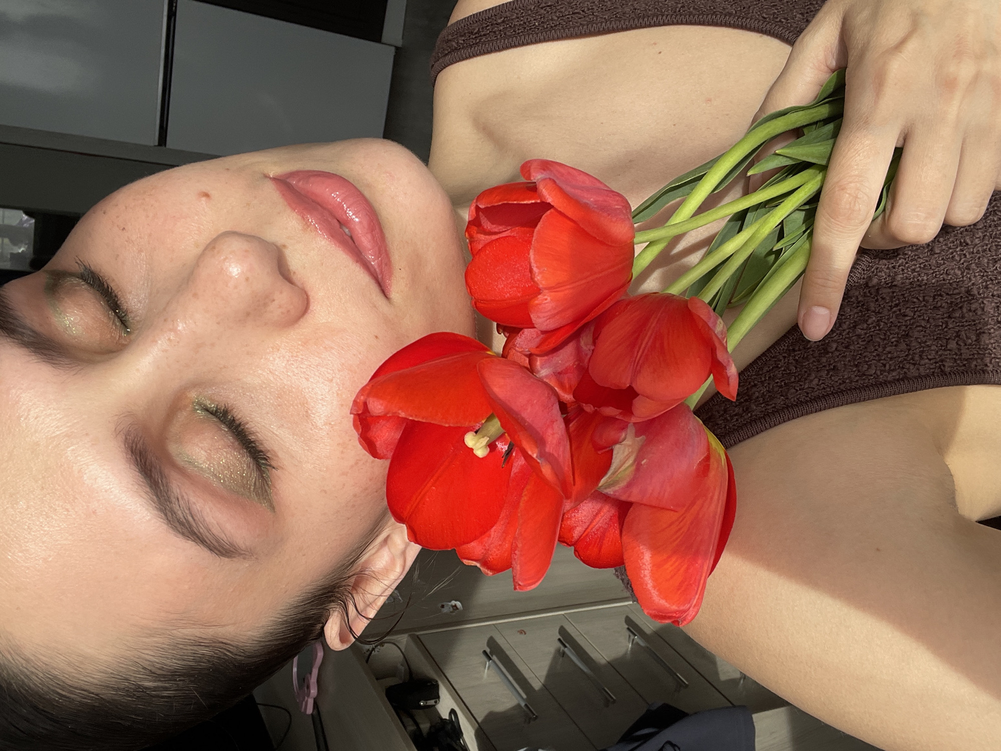 A woman is lying on her side, with a bouquet of red tulips resting on her chest and shoulder. She is wearing a brown top. Her eyes are closed and she has makeup on her eyes and lips. The background appears to be a room with a cabinet. There is no verifiable information to pinpoint the location to Izhevsk, Russia.