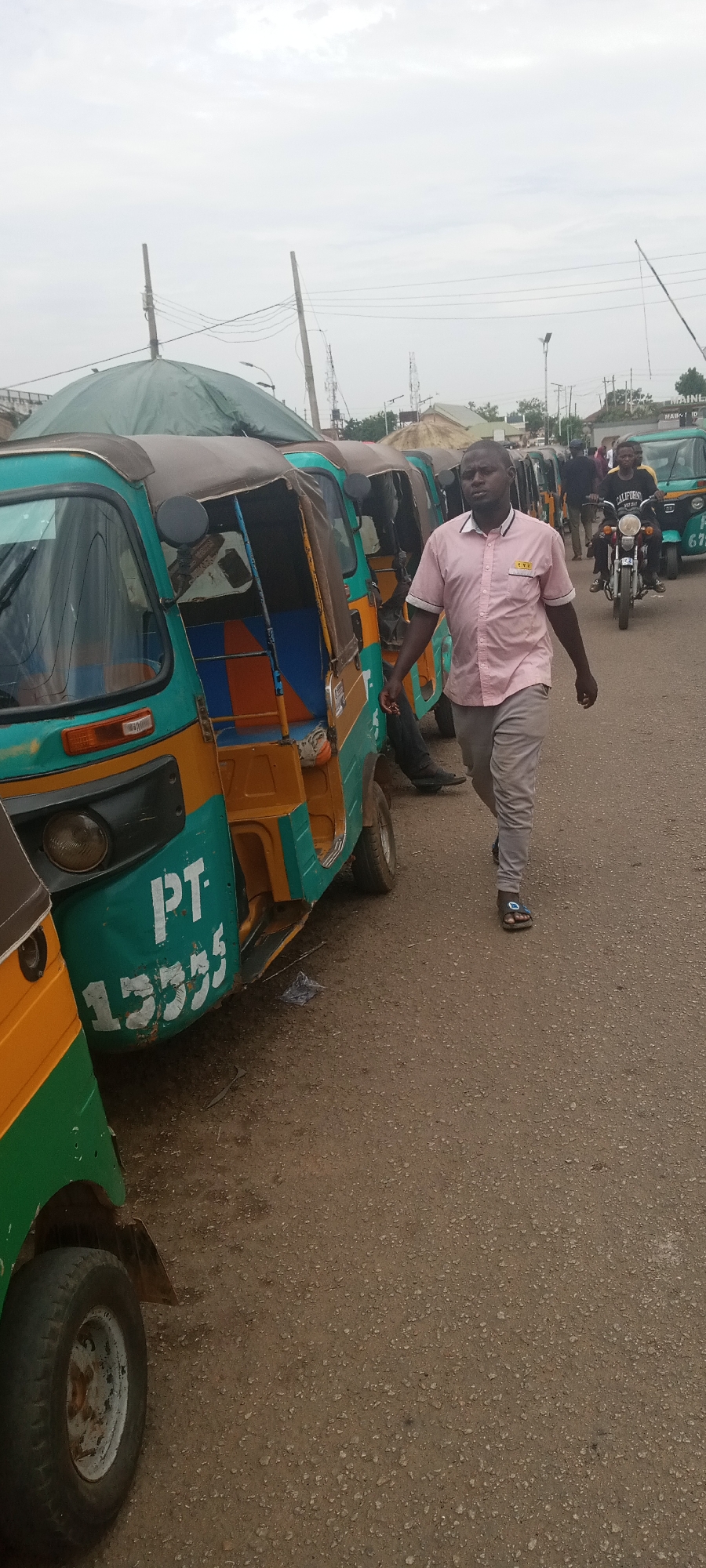 The image shows a row of auto rickshaws parked on a street in an urban area. A man in a pink shirt and gray pants is walking past them. In the background, a motorcyclist rides past and people mill about. The road surface is dusty and gray. The sky is overcast. The text "PT 13335" is prominently displayed on the side of one of the auto rickshaws. The location is described as Unknown, Unknown.