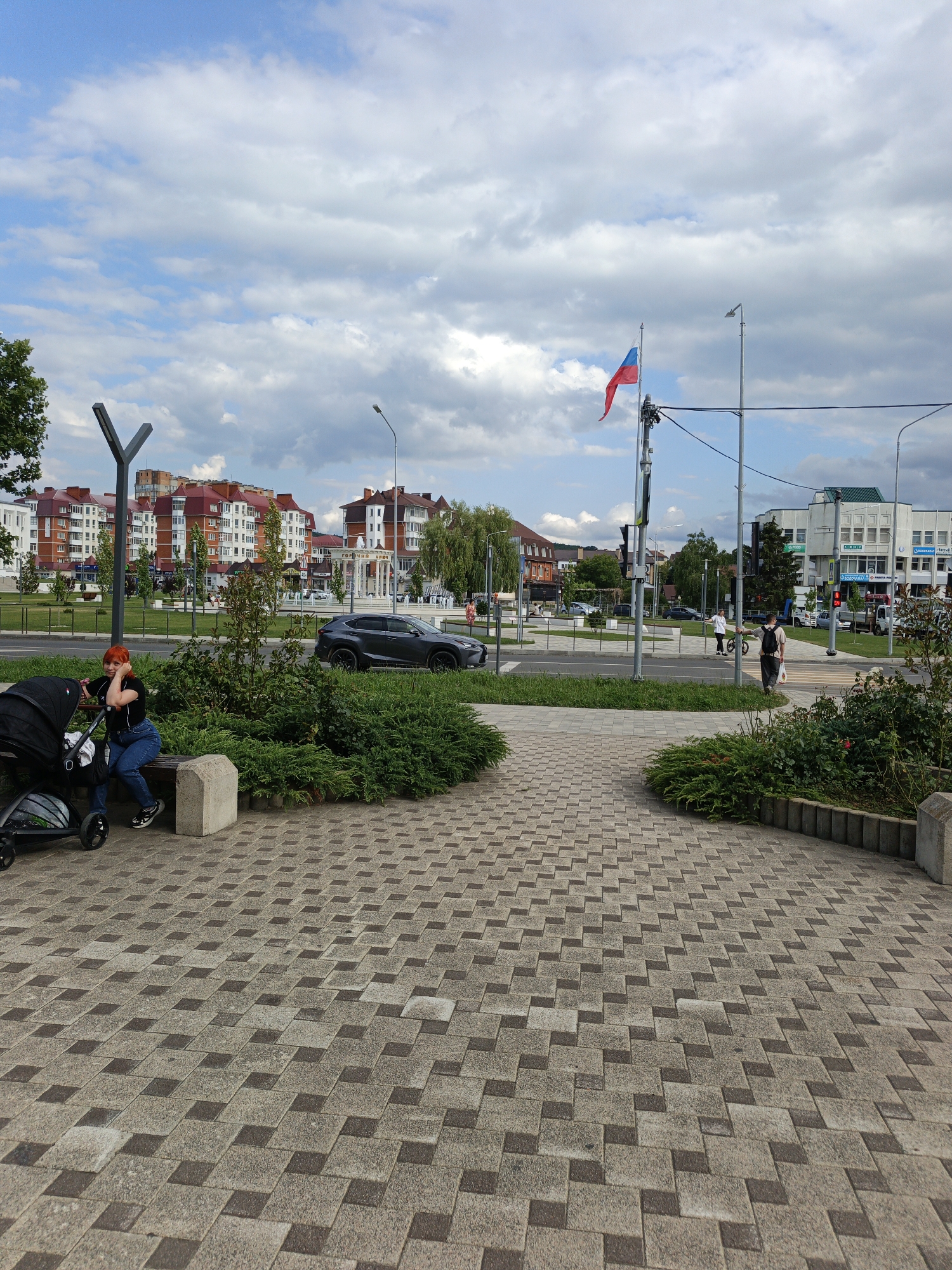 This image captures a street scene in Goryachiy Klyuch, Russia, on a partly cloudy day. In the foreground, a woman with vibrant red hair sits on a bench next to a black stroller. She is dressed in a black t-shirt and blue jeans and appears to be resting, leaning her head on her hand. The bench is situated on a paved area with a distinctive geometric pattern of light and dark gray square tiles.
Behind the woman, a neatly trimmed green hedge separates the pedestrian area from a road. A dark gray Lexus SUV is parked or driving on this road, with other vehicles and buildings visible in the background. The buildings are mostly residential, characterized by red-tiled roofs and light-colored facades. A Russian flag flies from a flagpole in the middle distance, adding a patriotic element to the scene.
Further down the street, people can be seen walking and crossing the road at a pedestrian crossing. Traffic lights and streetlights are also visible. The overall atmosphere seems calm and ordinary, depicting a typical day in a town. The time of day appears to be late afternoon or early evening, given the lighting and cloud cover. There is no legible text clearly visible in the image.