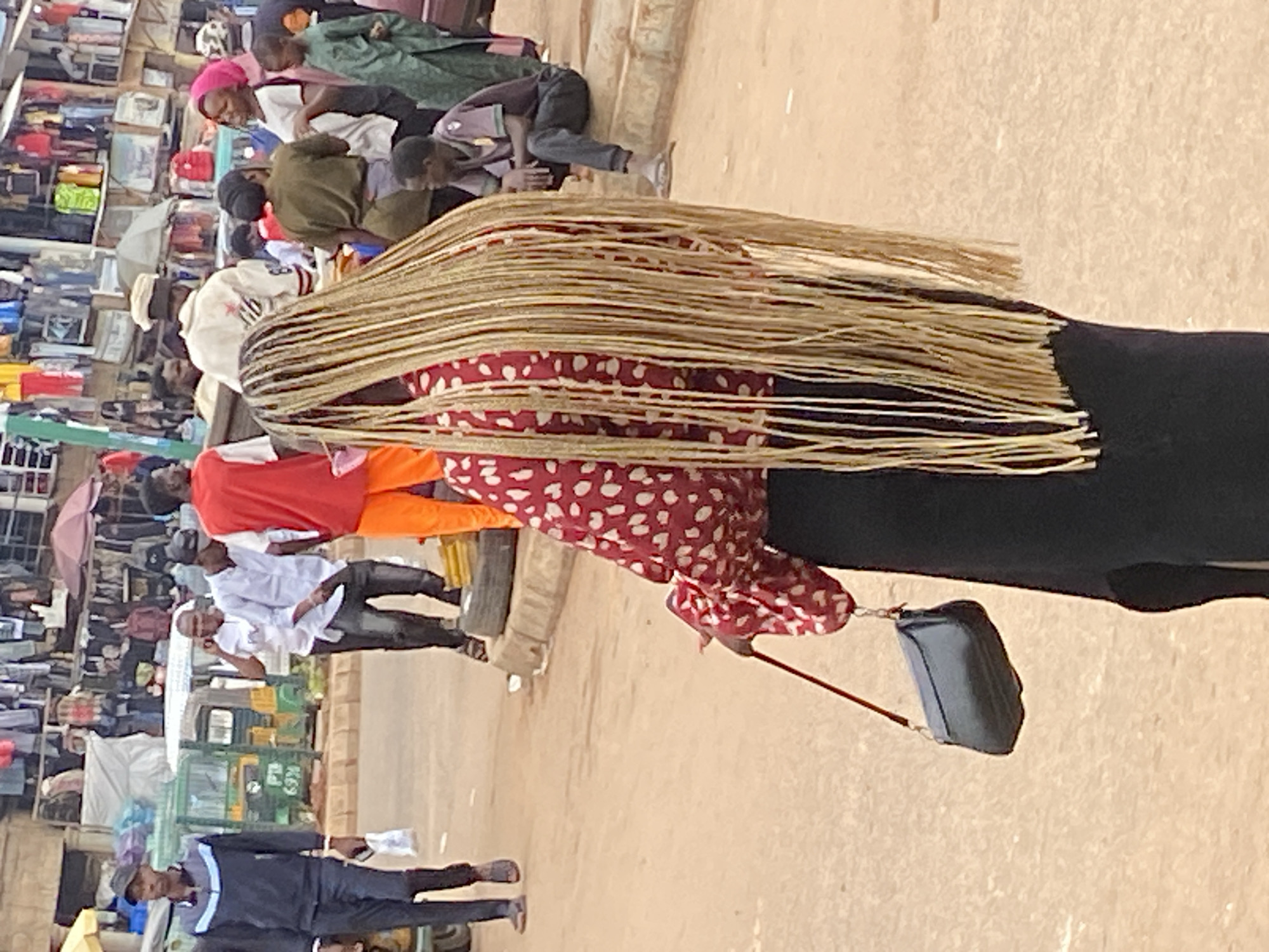 The image depicts a bustling marketplace scene in Bukuru, Nigeria. The central subject is a woman with long, blonde braided hair, viewed from behind. She is wearing a red top with a white polka-dot pattern and a long black skirt. A dark-colored purse with a red strap hangs from her shoulder.
Several other individuals are visible in the background. To the left, a man in a dark tracksuit with blue stripes walks away from the camera, carrying a white plastic bag. In the middle ground, other people are engaged in various activities; one person is on a phone, and others are walking or standing. A green motorized tricycle, a common mode of transport in the area, is parked on the left side of the image.
The marketplace environment is evident with stalls displaying clothing and goods, suggesting a commercial area. The ground is unpaved, typical of many Nigerian markets. The overall impression is one of everyday life and commerce in a Nigerian urban setting.