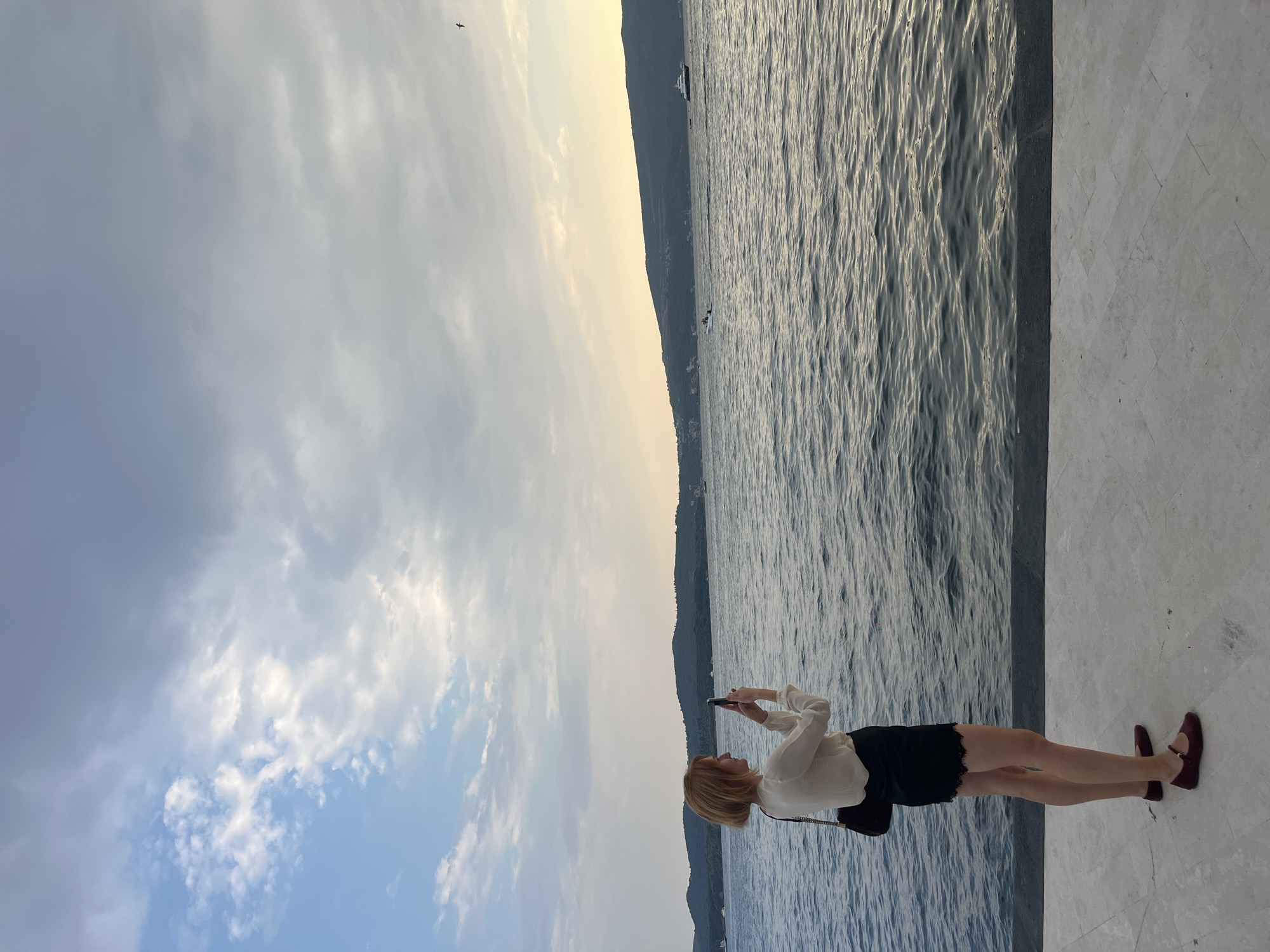 A side profile of a person with short blonde hair, wearing a cream-colored long-sleeved blouse, a black lace-trimmed skirt, and burgundy flats, stands on a paved walkway next to a body of water, holding up a smartphone to take a picture. The person is positioned on the left side of the frame, facing right. Behind them, a calm, grey-blue body of water stretches to a distant, tree-covered shoreline under a cloudy sky. The sky is a mix of grey and white clouds, with a hint of sunlight breaking through on the right horizon. Several boats, including a white yacht, are visible on the water in the distance. The walkway in the foreground is made of large, light-colored square tiles.