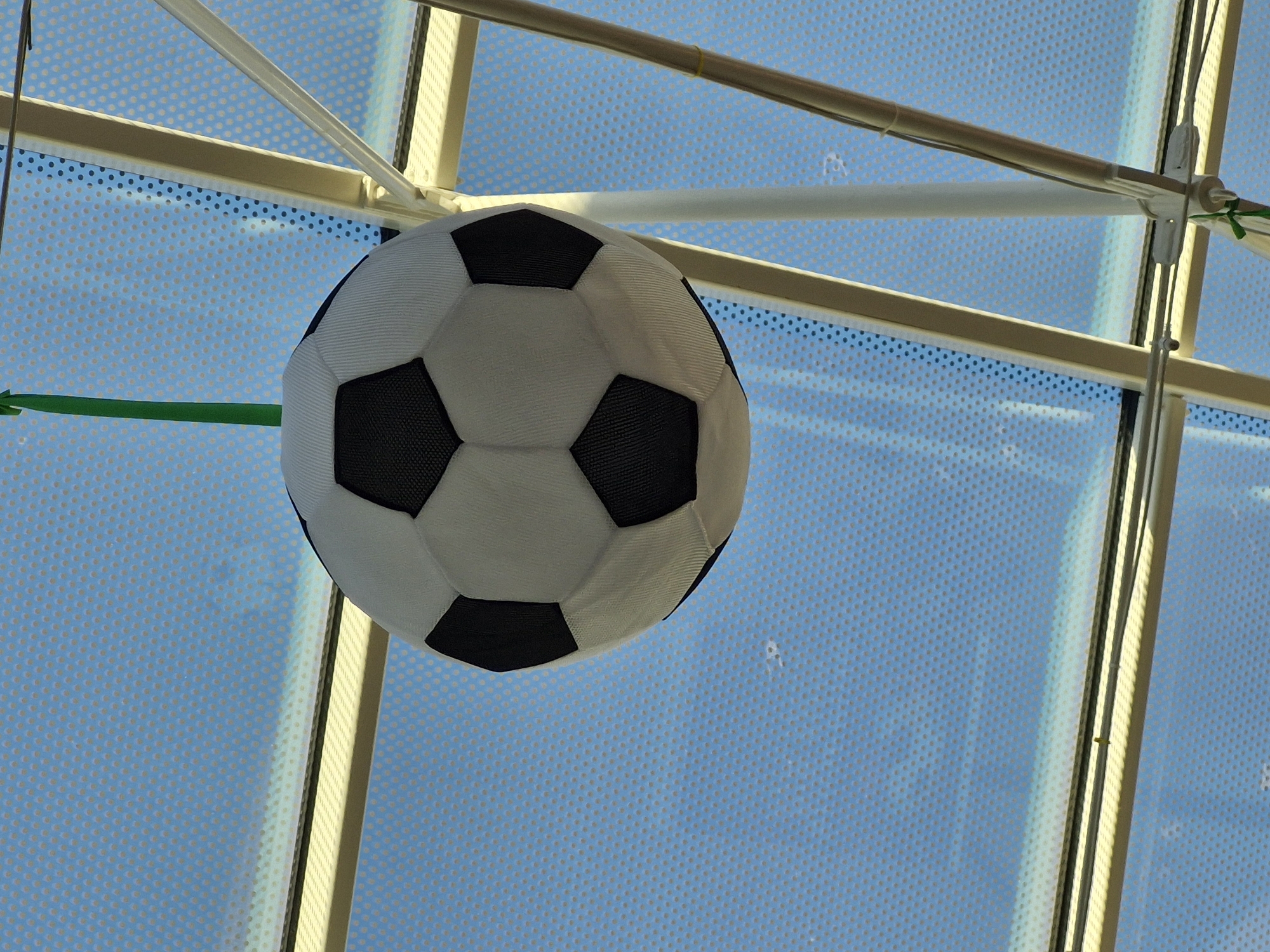 A black and white soccer ball hangs from a green string from an off-white ceiling structure. The ceiling is made of light tan beams with a blue patterned background visible through some of the panels. The soccer ball is partially obscured by the ceiling structure. The scene is brightly lit, suggesting an outdoor or well-lit indoor environment. This image does not provide any identifiable landmarks or features to suggest a specific location within the city of Bremen, Germany.