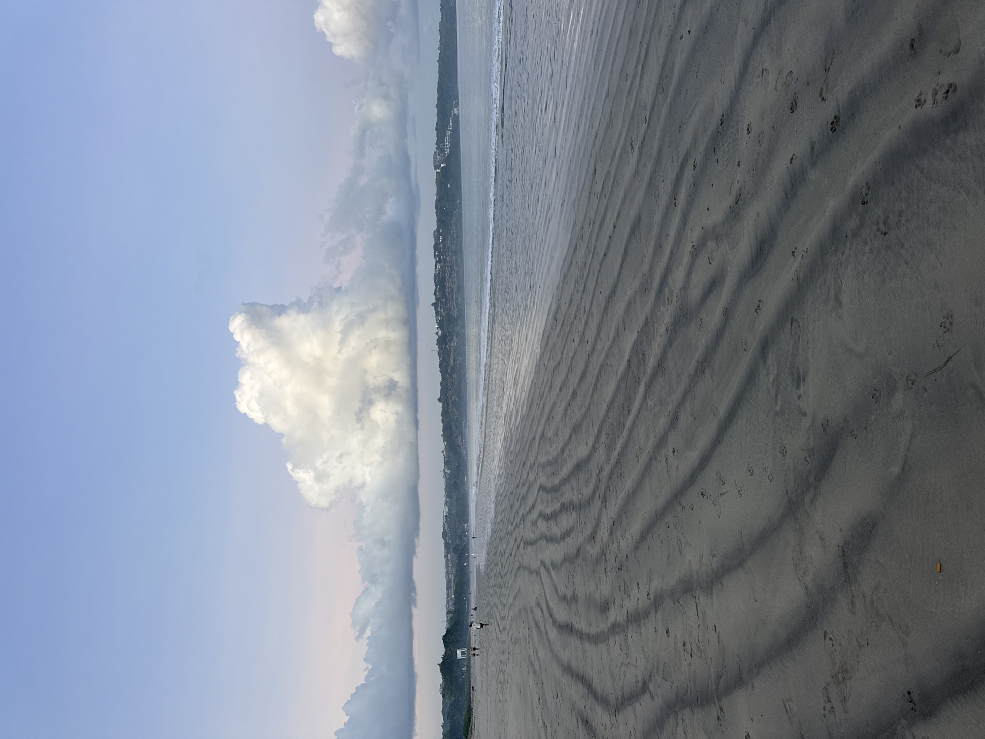 The image depicts a beach in Kuta Selatan, Indonesia. The scene is dominated by a wide sandy beach marked with wave patterns and scattered footprints. The ocean meets the sand in a shallow wave line. In the distance, a coastline with buildings and vegetation is visible. The sky is partially cloudy, with a large cumulus cloud formation on the left side of the frame. Two small figures of people are walking on the beach in the mid-ground.