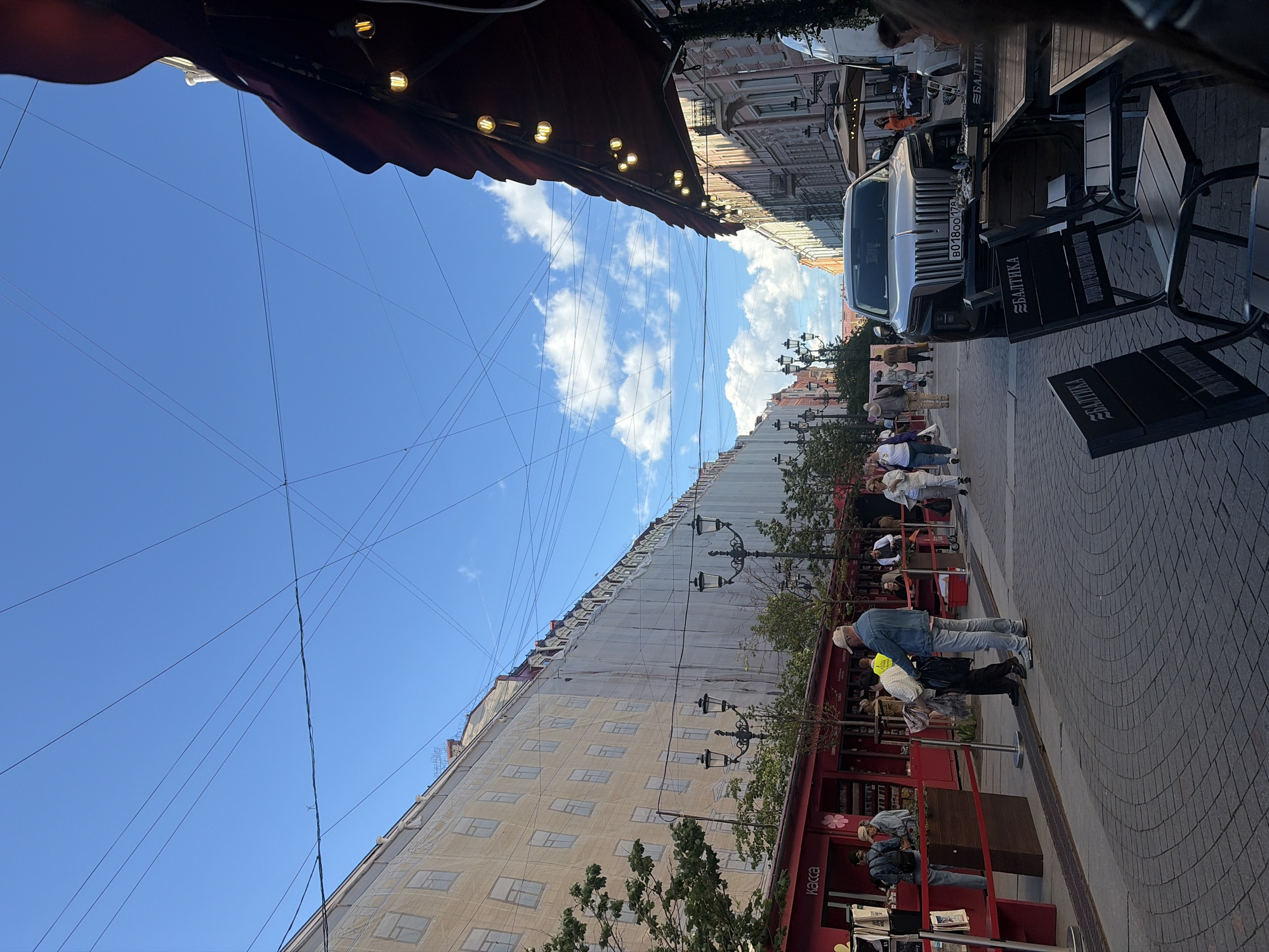 An outdoor street scene shows a wide, cobblestone-paved street in front of shops and restaurants under a bright blue sky with scattered clouds and overhead wires. On the left, a long building is partially covered with a large white tarp, suggesting ongoing renovation or construction, with windows visible through the covering. Red awnings and storefronts line the street on the left, with a sign that reads "Касса" (Kassa, meaning "cashier" or "ticket office"). Several people are walking on the sidewalk and in front of the shops.
In the center of the street, a large, silver luxury SUV with a prominent grille is parked. The license plate is visible.
On the right side of the street, a restaurant or cafe area with tables and chairs is set up under a dark red awning decorated with string lights. The chairs have the logo "БАЛТИКА" (Baltika) on them, which is a Russian brewing company. People are seated at some of the tables. Further down the street, more buildings and streetlights are visible, giving a sense of an urban environment. The overall impression is a bustling street on a sunny day, with a mix of shops, dining, and vehicular traffic.