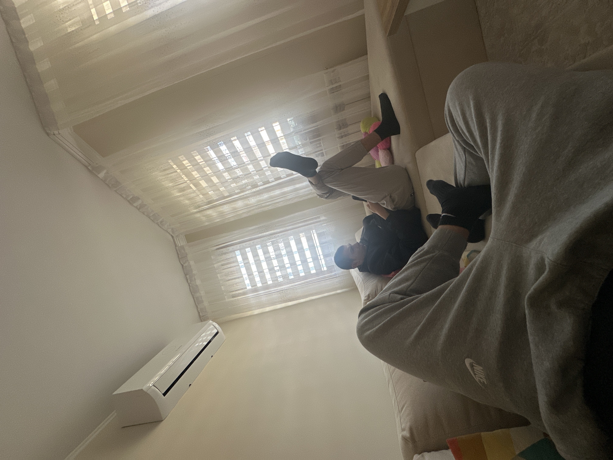 A man is lying on a couch in a room with blinds on the windows. Another person is sitting on the couch, partially obscuring the view. An air conditioner is mounted on the wall. The specific location within Plovdiv, Bulgaria, cannot be determined from the image.