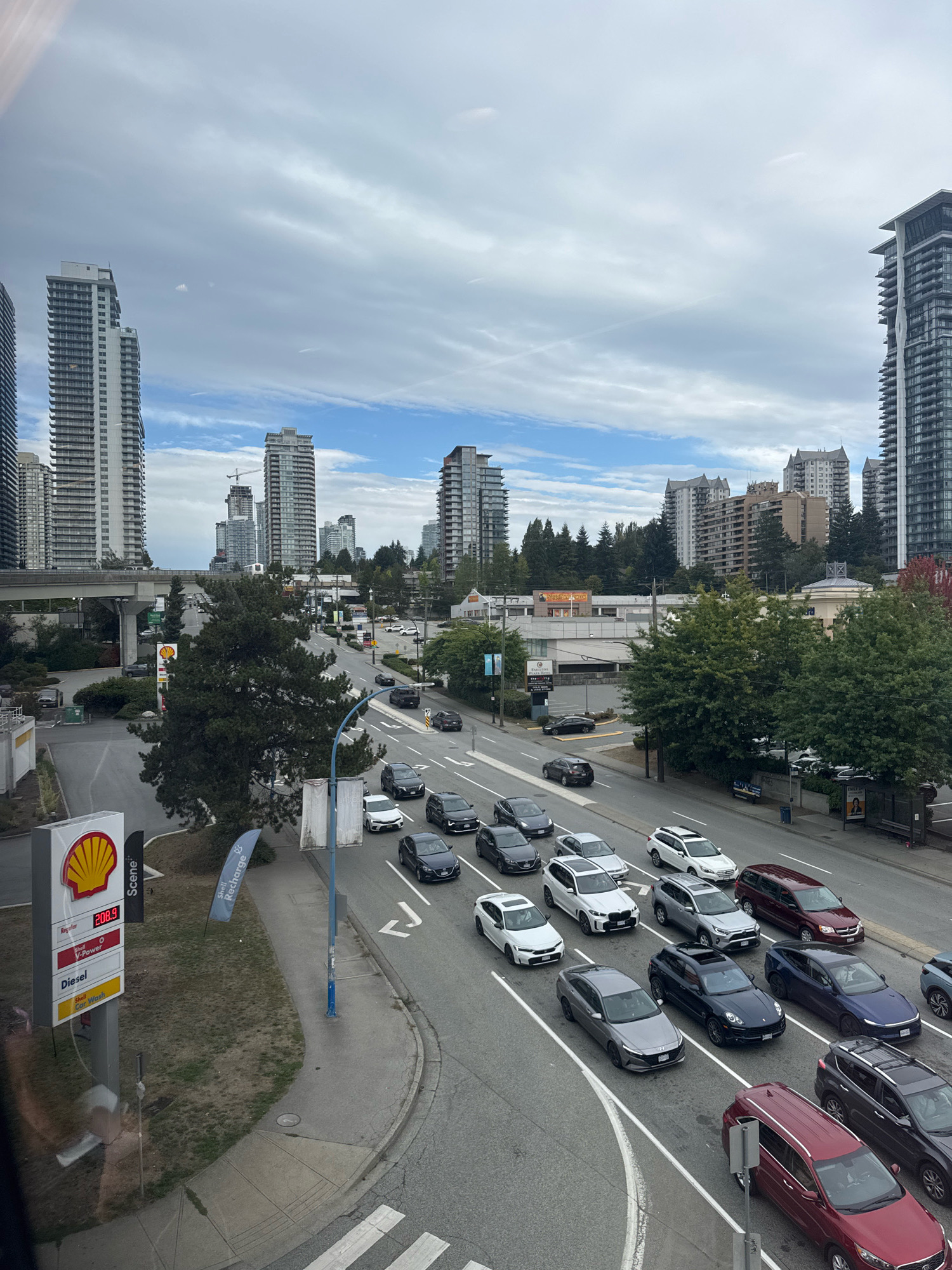 The image depicts an urban scene in Burnaby, Canada, likely taken from an elevated position looking down onto a multi-lane road with moderate traffic. Several tall, modern residential buildings dominate the skyline, with some under construction, indicated by a crane in the background. The sky is partly cloudy, suggesting an overcast or transitional weather day.
On the right side of the road, there are parked cars and a bus stop with a poster. The road itself shows white lane markings and a pedestrian crossing. Vehicles are proceeding in both directions, with a noticeable line of cars in the right lane of the visible traffic. Among the visible vehicles are a dark red minivan, a dark blue SUV, a dark grey SUV, a white SUV, and several other cars of various colors and models.
On the left side of the road, there is a Shell gas station with a prominent red and yellow sign displaying fuel prices. Adjacent to the gas station, there is a grassy area and trees. A monorail track runs overhead on an elevated concrete structure. The overall impression is a busy, contemporary urban environment characterized by high-rise development and vehicular activity. There are no identifiable people in the image.