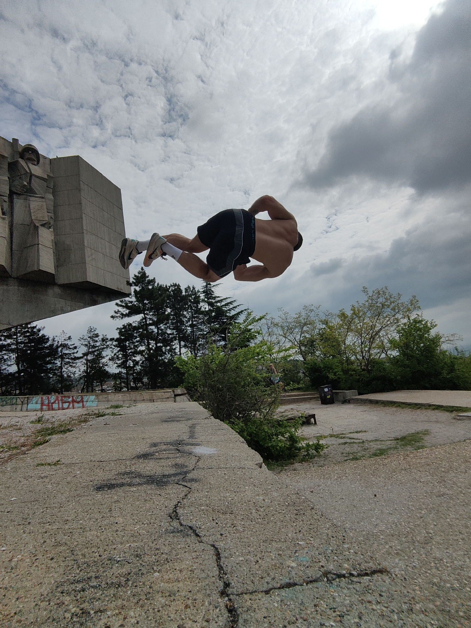 A man is captured mid-air in a dynamic pose. He is shirtless, wearing dark shorts and sneakers with white socks. He appears to be performing a parkour move or a jump. He is in the foreground.
In the background, there is a large concrete monument with figures, and behind that are trees, the sky, and clouds.
The setting is an urban area, appearing to be a park or a public space with paved areas. There are cracks in the pavement and some graffiti on a wall near the monument. This is the Pantheon, a communist era monument, located in Varna, Bulgaria.