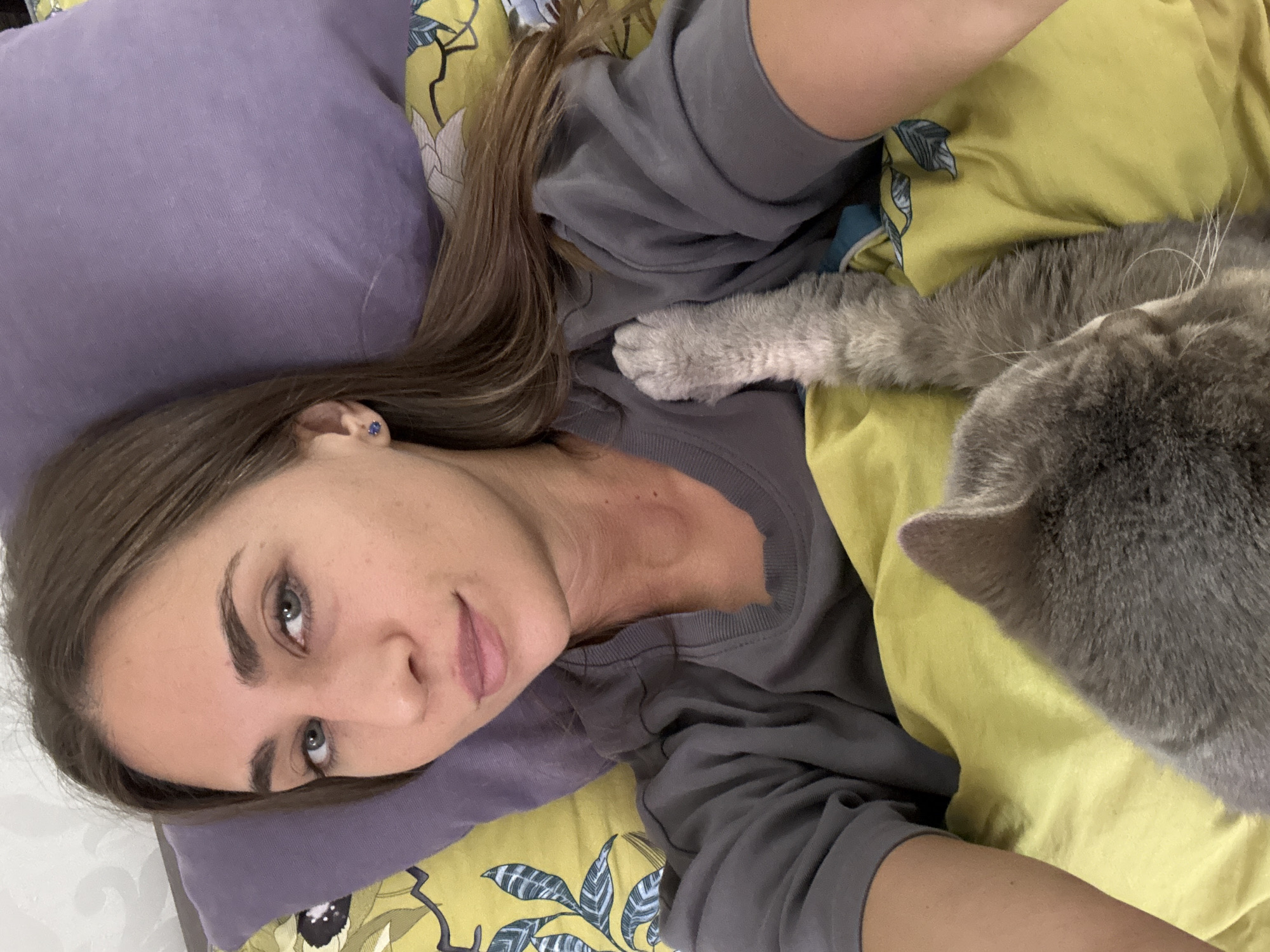 A woman is lying on a bed with a gray cat. The woman has light brown hair and is wearing a gray shirt. She is resting her head on a purple pillow. The bed has a yellow floral patterned sheet. The cat is lying on the sheet next to the woman, with its paw on her chest. The photo appears to have been taken from above. The location of the photo is unknown, and there is no specific indication that it was taken in Zanevka, Russia.