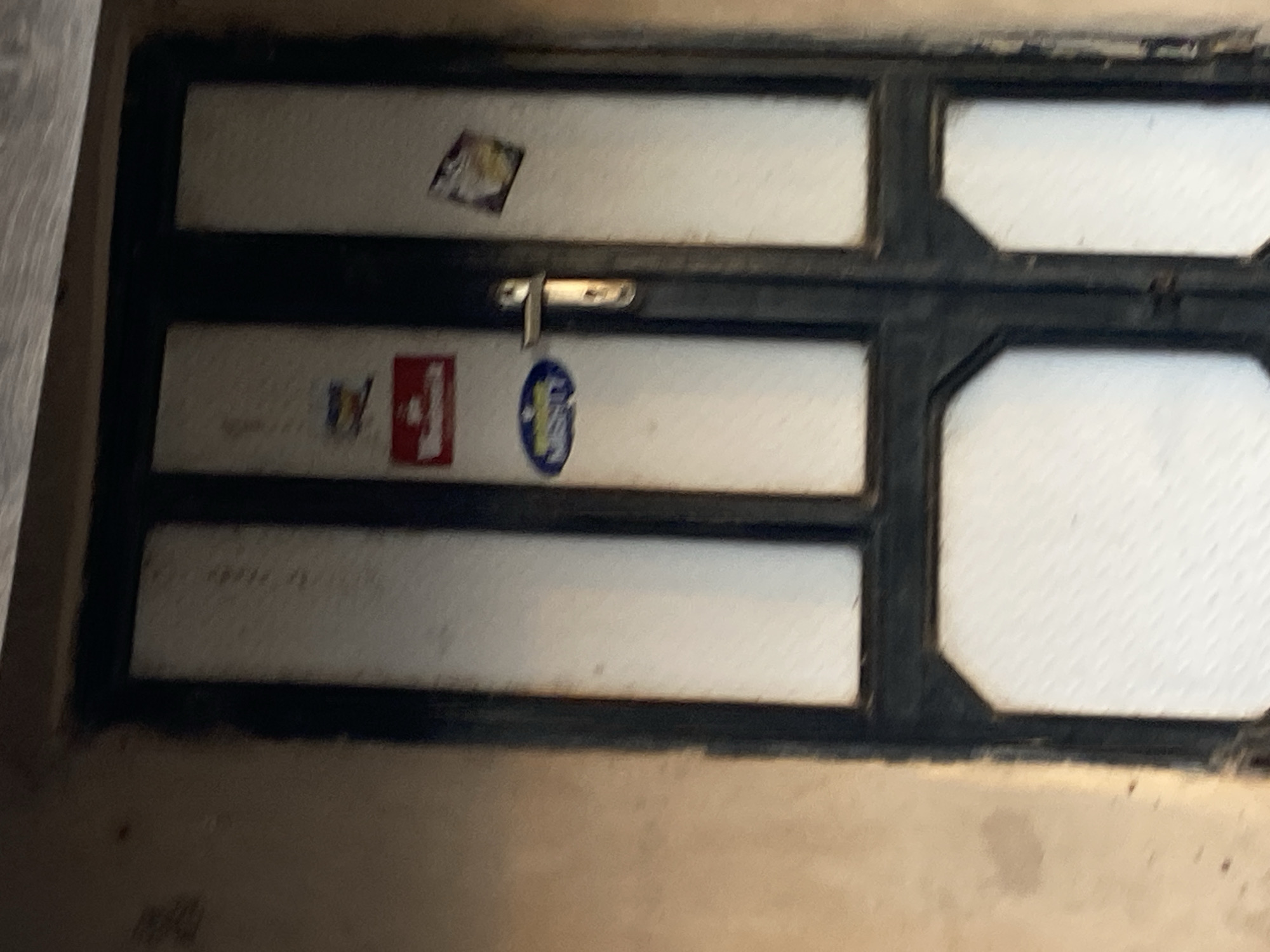 The image shows a close-up, slightly blurry view of a dark-colored door with glass panes. The door appears to be old and weathered. There are several stickers on the door, including a red rectangular one with white lettering, a blue oval sticker with white lettering, and a smaller, colorful sticker. A silver door handle is visible on the right door. The wall next to the door is a muted tan color. The scene appears to be an exterior shot, possibly in an urban or semi-urban environment. The location of Jauro Yinu, Nigeria is not visually discernible from the image itself.