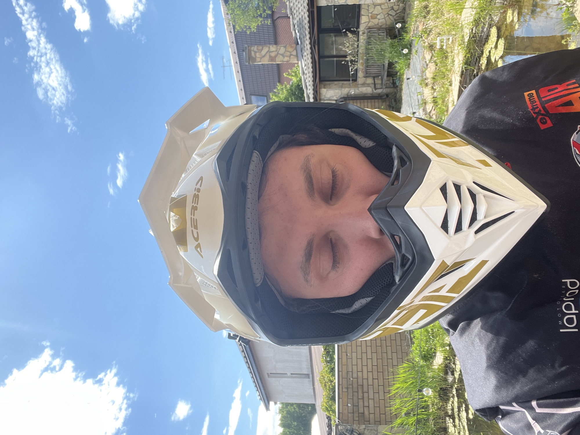 A close-up, waist-up, vertical photograph depicts an individual wearing a white and gold motocross-style helmet. The individual, whose eyes are closed, wears a black long-sleeved shirt displaying "laplad MOTO WEAR" text on the lower left and "eXtreme AIR" text on the lower right. The helmet features the "ACERBIS" brand logo on the top visor and gold graphic designs on its sides. The background reveals a sunny outdoor residential setting. To the left, a light-colored brick wall, green foliage, and a two-story building with a dark roof are visible. To the right, a pond with green surface vegetation, green grass, a stone-clad house with a dark tiled roof and stone chimney, and a wooden bench are present. The sky above is blue with scattered white clouds. The scene is located in Ореховка, Russia.