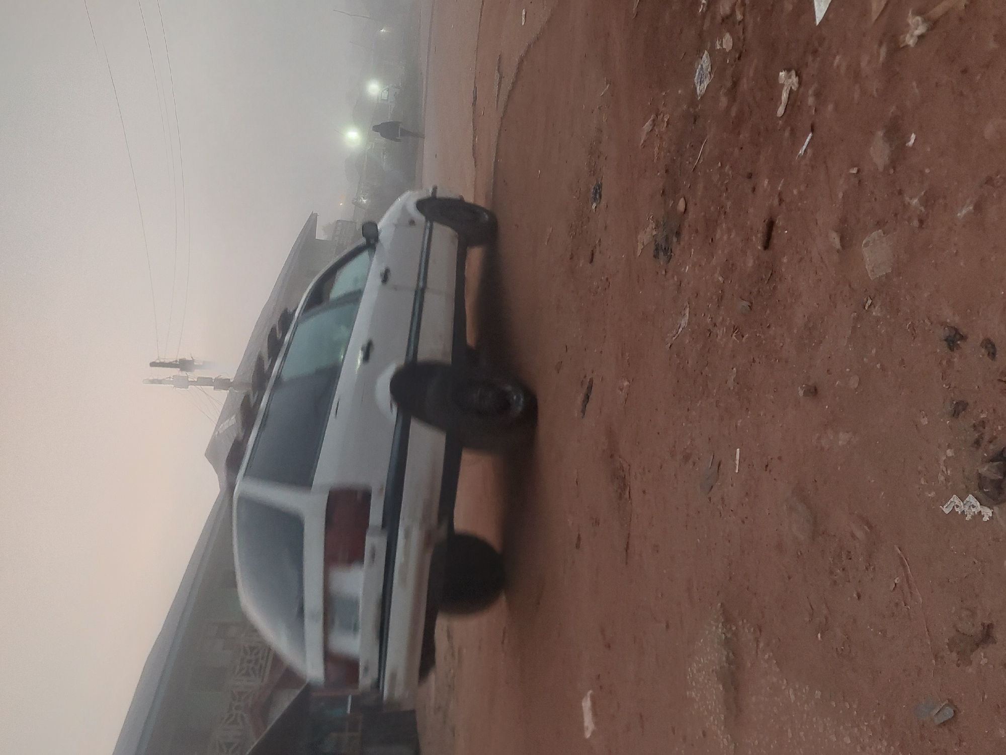 The image depicts a foggy, early morning scene in Bukuru, Nigeria, with a white station wagon driving on a dirt road. The car's rear is visible, with its tires and suspension appearing raised, suggesting it might be an off-road modification or is experiencing mechanical issues. The background is obscured by dense fog, with faint outlines of buildings and utility poles visible. A lone figure is seen walking in the distance, partially illuminated by streetlights. The ground is covered in reddish-brown dirt with scattered debris, indicating a rural or less developed area. The overall atmosphere is muted due to the fog and low light conditions.