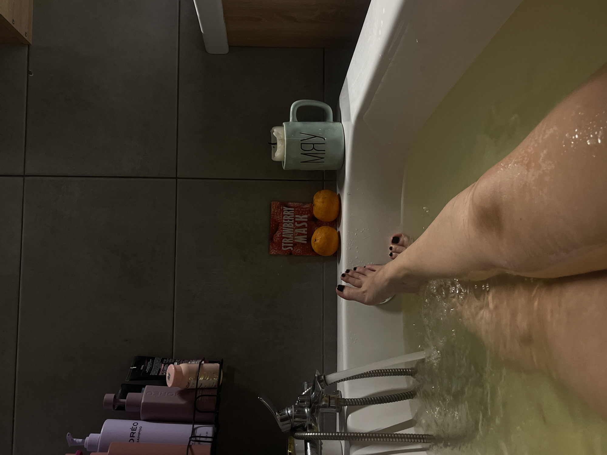 A person is taking a bath. The lower half of their body is submerged in water, with their feet visible near the edge of the tub. Two oranges, a mug with the word "MAY" printed on it, and a "STRAWBERRY MASK" package are placed on the ledge of the bathtub. A rack of shampoo and conditioner bottles is mounted on the wall next to the tub. The location appears to be a bathroom. There is no evidence to suggest this is located in Мосметаллоконструкция Онт, Russia.