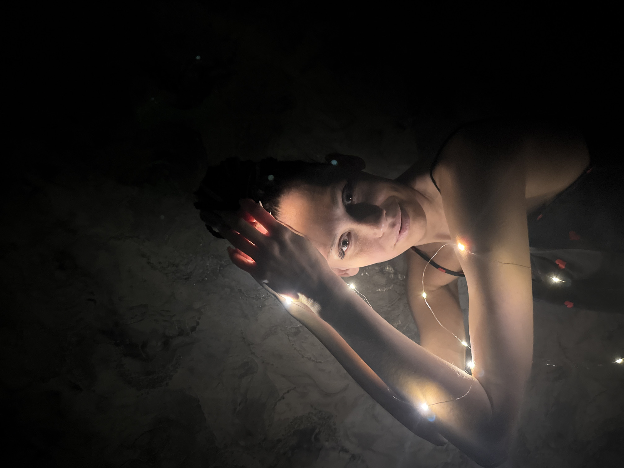 A woman is lying in water, possibly a bathtub or shallow pool, in a dimly lit environment. She is positioned at an angle, looking towards the camera with a slight smile. Fairy lights are draped across her arm and around her, casting a warm glow on her skin and the surrounding water. The background is dark and indistinct, with marble-like textures visible in the water or the surface below. The image is suggestive of a relaxing or intimate scene. There is no information available about this being in Rusanovo, Russia, in the image.