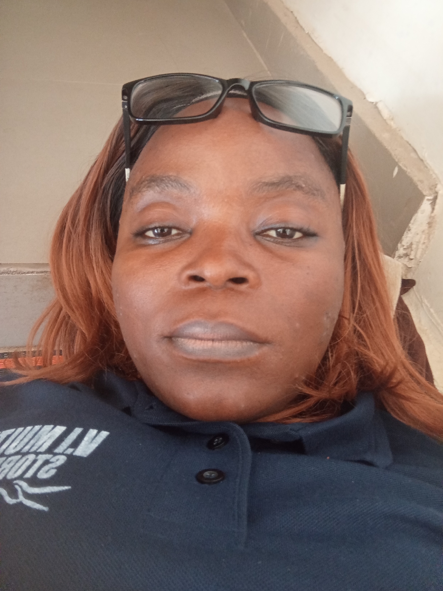 The image is a close-up, waist-up selfie of a dark-skinned woman with reddish-brown hair and glasses perched on her head. She is wearing a dark blue polo shirt with a white graphic design on the left side of the chest, which appears to be text and a bird-like illustration. The background is a plain, light-colored wall with some texture and discoloration, suggesting an indoor environment, possibly a room in a residential or commercial building. The lighting is natural and appears to be coming from the front, casting soft shadows on her face. The overall impression is of a casual, personal photograph. The image does not provide any specific visual cues that would allow for the precise identification of its location within Jalingo, Nigeria.