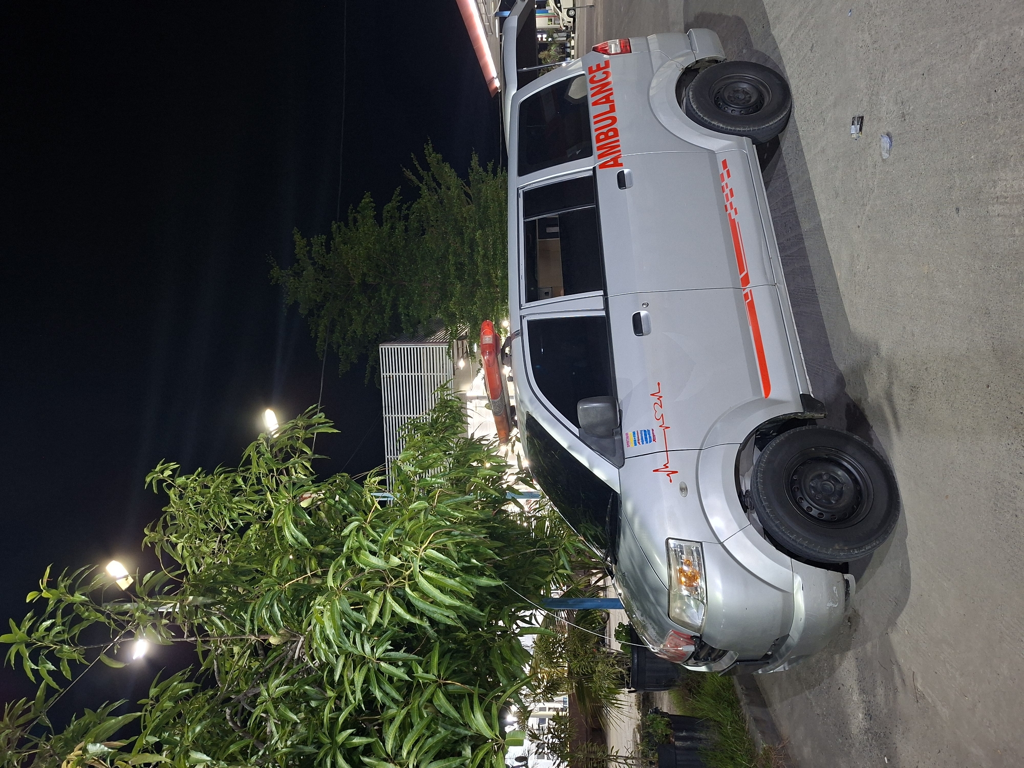 The image captures an ambulance parked at night, likely in Keuniree, Indonesia, given the context. The ambulance is silver with red lettering spelling out "AMBULANCE" on its side. It features a red emergency light bar on its roof and a red heartbeat line graphic with a heart emblem on the front fender. The vehicle is positioned on a paved surface, possibly a parking lot or road, with a large tree with green leaves to its left. In the background, there's a building with horizontal white louvers and some lighting. The scene is illuminated by artificial lights, creating shadows and highlighting the ambulance.