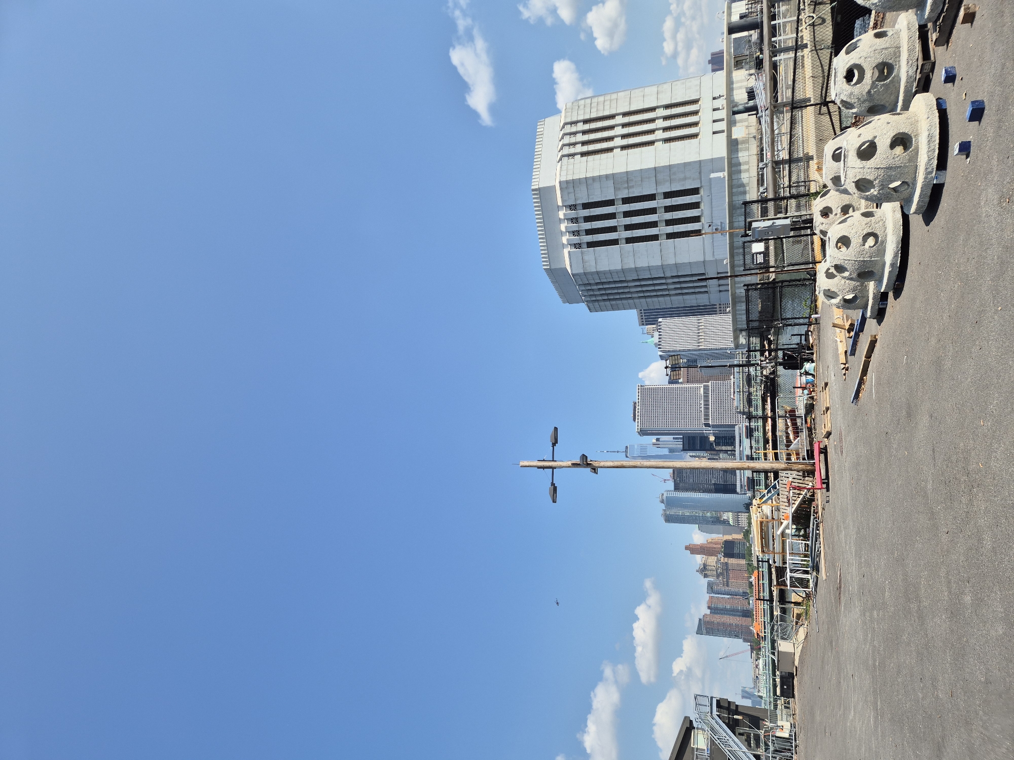 This is a wide-angle, eye-level, full outdoor shot of a city waterfront on a clear, sunny day. The foreground is an asphalt-paved area with several large, light-colored, dome-shaped concrete structures arranged in a row. These structures appear to be some type of artificial reef or breakwater units, each with multiple circular openings. Behind these concrete forms, a black chain-link fence runs horizontally across the mid-ground, separating the paved area from a body of water. Beyond the fence, various construction materials, including metal scaffolding and wooden planks, are visible.
In the background, a dense skyline of New York City skyscrapers dominates the horizon. Notably, the iconic One World Trade Center is identifiable among the buildings. A large, light-colored, modern building with vertical window elements stands prominently on the right side of the image, close to the waterfront. A wooden pole with two modern streetlights is positioned in the middle ground, with a small aircraft visible in the sky to the left of the pole. The sky is a vibrant blue with scattered white clouds. The overall impression is one of urban development and waterfront activity in New York City.