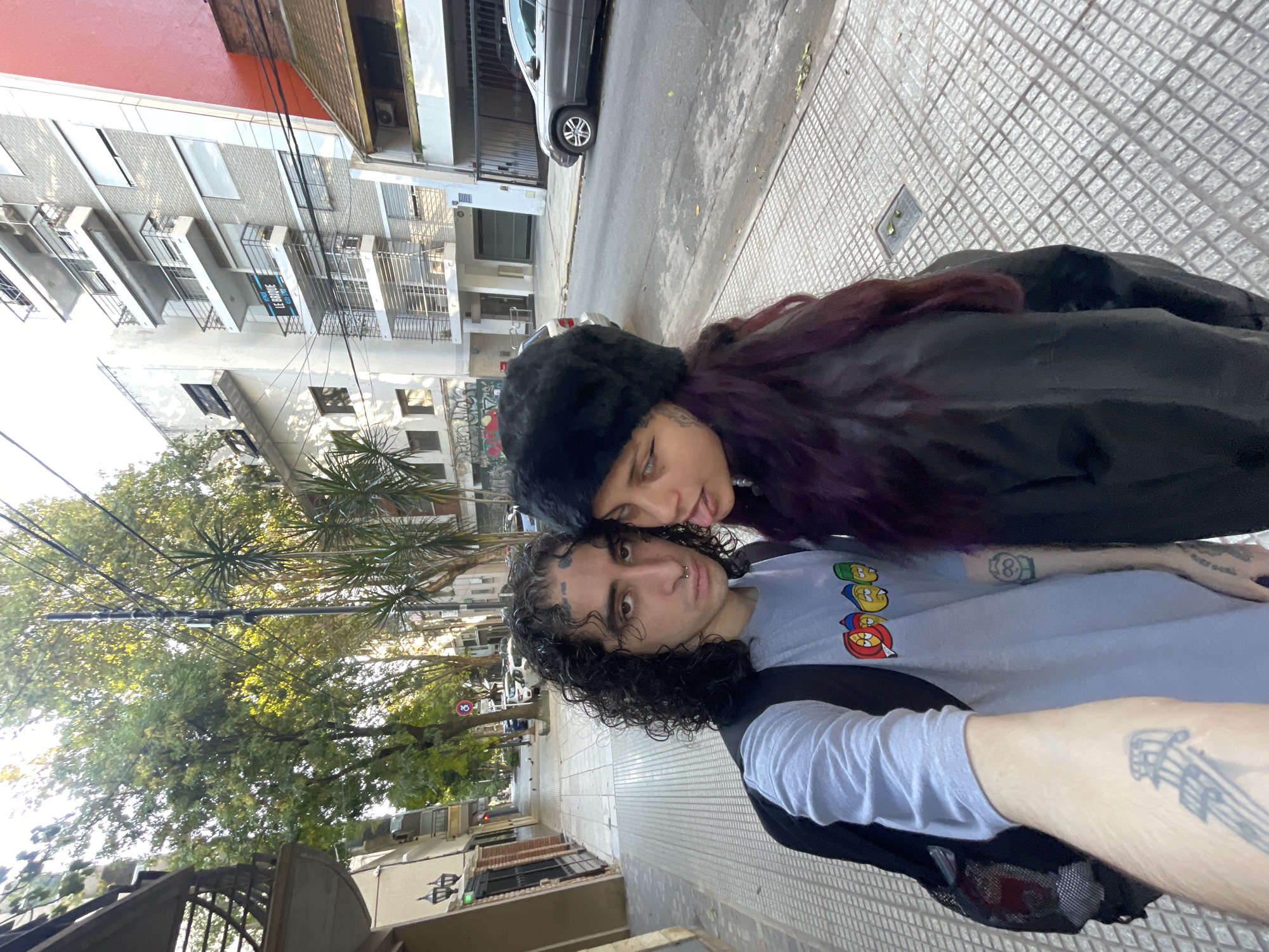 A selfie taken at street level in Buenos Aires, Argentina, shows a young couple. The man has dark curly hair, a nose ring, eyebrow tattoos, and is wearing a light grey t-shirt with cartoon characters and a black jacket. He has tattoos on his arms. The woman, wearing a black hat and a black jacket, has dark hair with a purple hue. The pair appear to be posing in a somewhat affectionate manner. The background reveals a typical urban street scene of Buenos Aires with buildings, trees, parked cars, and patterned sidewalks.