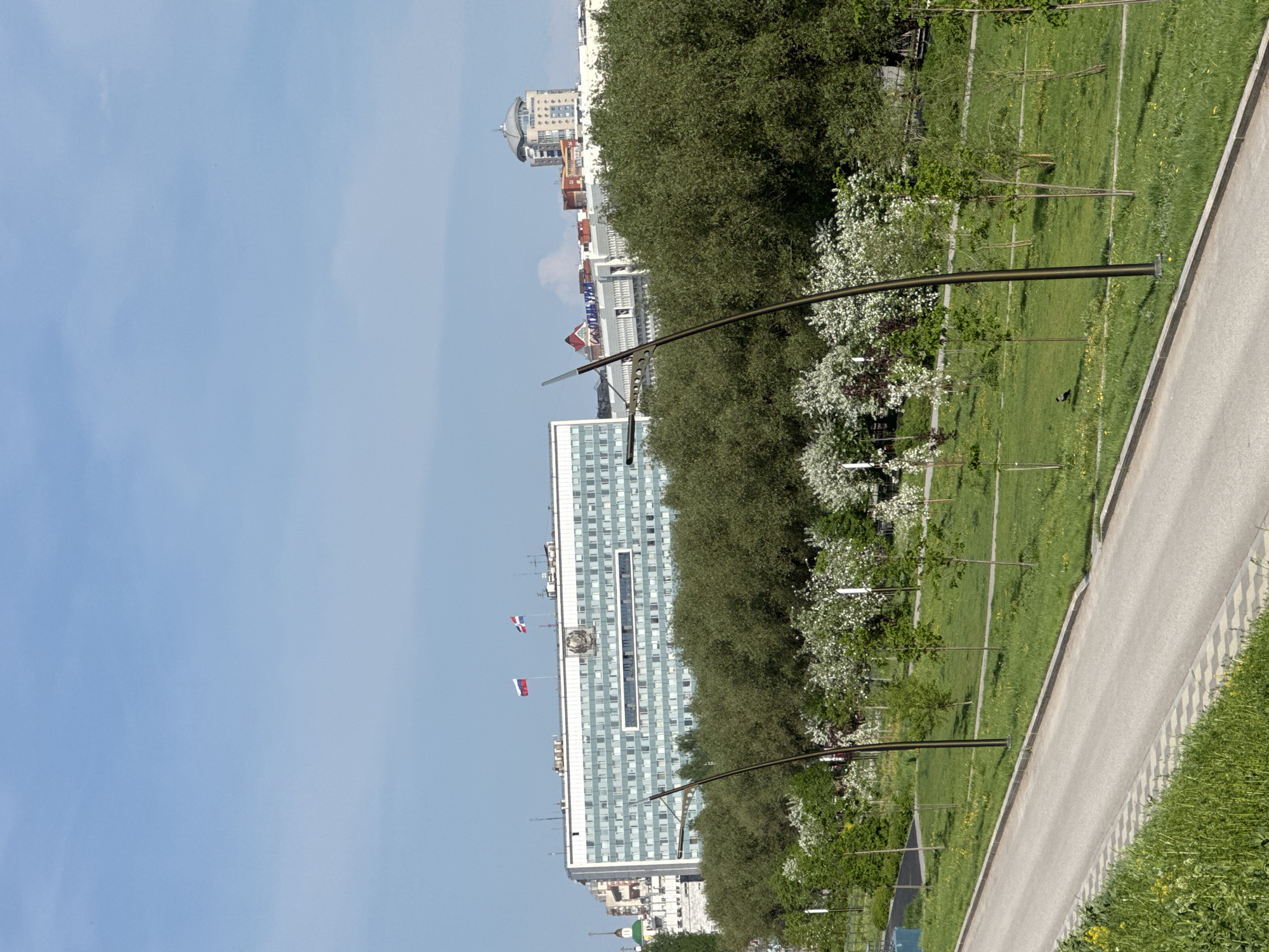 The scene presents a long, gray-paved pathway bordered on the right by green vegetation and on the left by a vast green lawn dotted with trees, some of which are flowering with white blossoms. Modern streetlights curve over the path.
Beyond the trees, a multi-story building dominates the skyline, its façade a grid of windows. Flags are mounted on its roof. Further in the distance, additional buildings and structures rise up.
The picture is taken from an elevated perspective, possibly from a high-rise building or a hilltop. The location is Perm, Russia, evidenced by the building which houses the regional government offices of the Perm Krai. The modern city landscape includes government buildings and park areas.