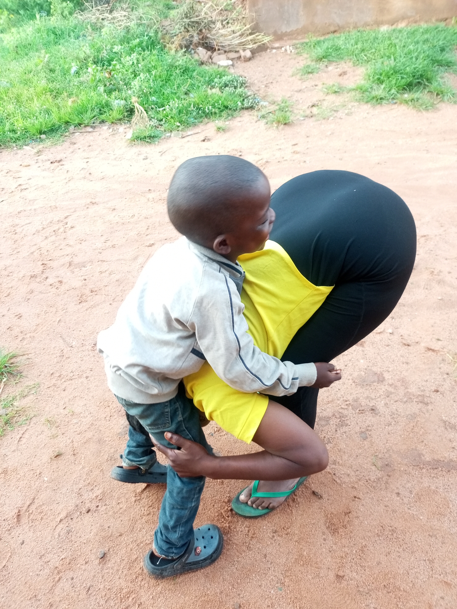 In the city of Tempure, Nigeria, a child wearing a light-colored jacket, blue jeans, and dark clogs is being held by a person wearing a yellow shirt and black leggings. The child's arms are around the other person's neck. They are standing on a dirt path with dry grass and some greenery in the background.