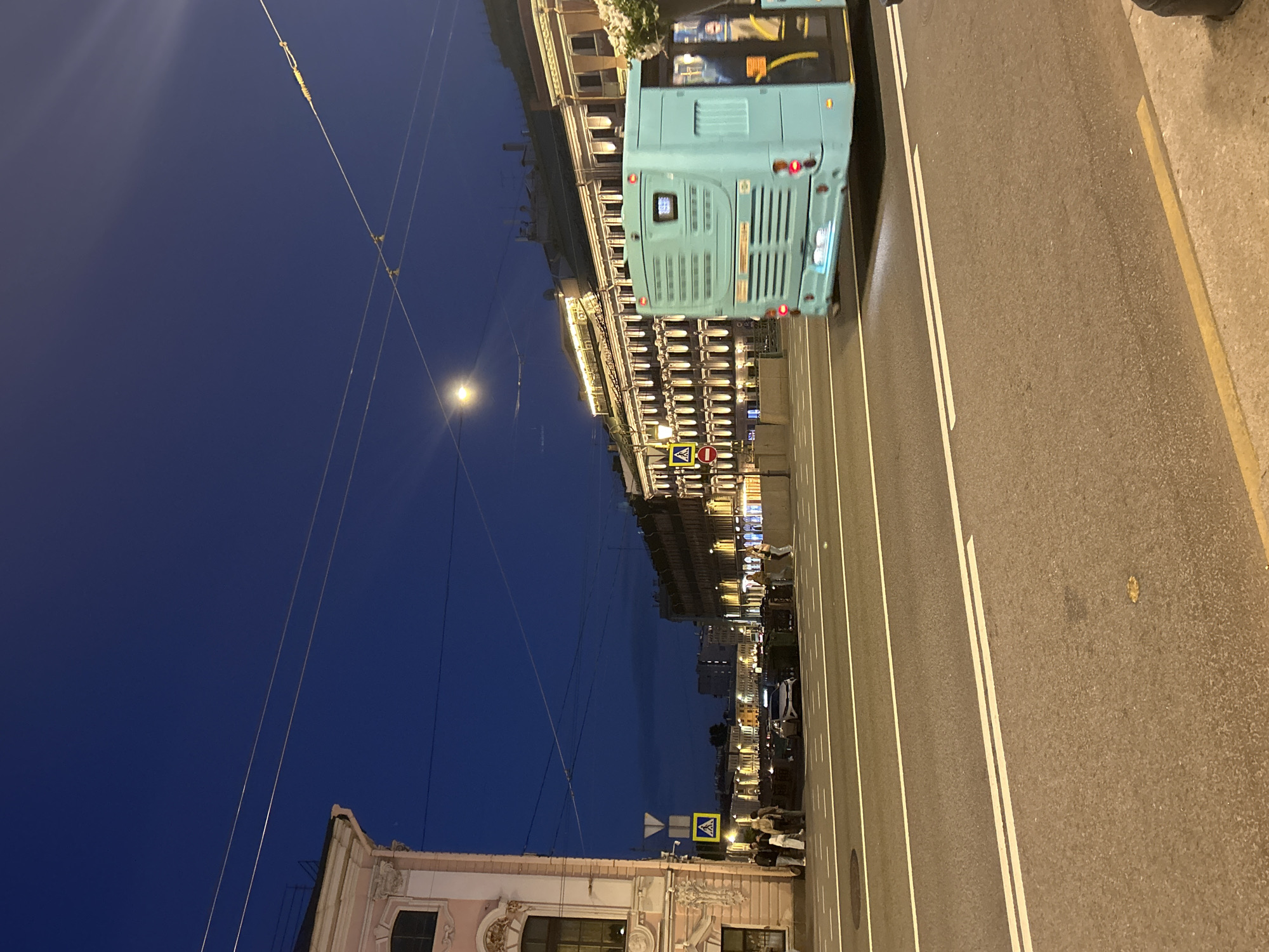 The image captures a street scene at dusk or night in an urban environment. A light blue bus is moving away from the camera, occupying the right half of the frame. The bus has its brake lights illuminated and displays license plates. In the background, a long building with numerous illuminated windows lines the street, suggesting it is a place of business or residences. Across the street from this building, another building with ornate architecture and decorative lighting is visible on the left side of the frame. Pedestrians are present on the sidewalk in front of the ornate building, and a car is parked further down the street. Traffic lines are painted on the asphalt road. Overhead, power lines crisscross the dark blue sky, and a bright moon or streetlamp is visible in the upper portion of the image.