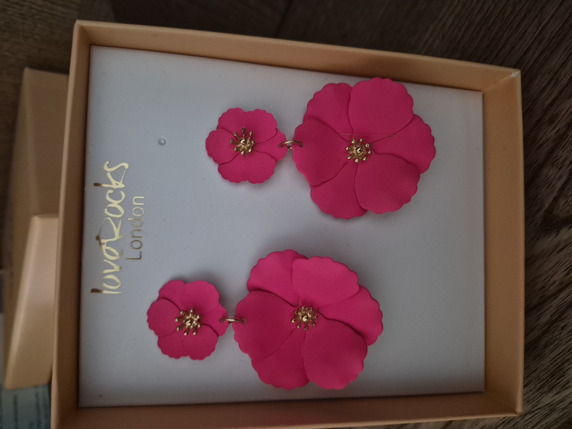 A pair of bright pink, flower-shaped dangle earrings are displayed in a tan box with the gold embossed text "love rocks London" on the lid. The earrings are made of a matte material and feature a smaller flower connected to a larger flower by a gold loop. The center of each flower has a cluster of gold stamen. The earrings are positioned on a light grey backing within the box, which is open and angled towards the viewer. The box rests on a dark wooden surface.
