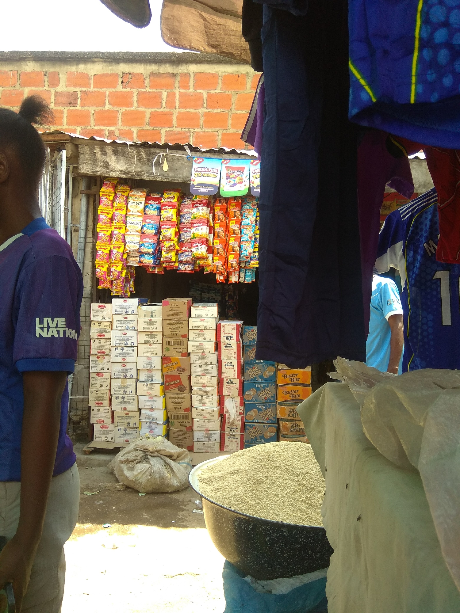 This image depicts a bustling market scene in Jalingo, Nigeria. In the foreground, a person wearing a purple t-shirt with "LIVE NATION" emblazoned on the sleeve stands to the left, partially obscuring the view of goods for sale. The market stall features an abundance of packaged snacks, including boxes of Kellogg's Cornflakes and other colorful bags. Behind the snacks, stacks of what appear to be cartons of goods are neatly arranged.
Further back, a man in a light blue t-shirt is visible, suggesting ongoing commerce. The market is set against a backdrop of a brick wall and a corrugated metal roof, indicative of an outdoor or semi-enclosed marketplace. The overall impression is one of a vibrant local economy with a variety of products available. Visible text on the packaging includes "MEGA FUN 2X BIGGER!", "Popzilla", "CORNFLAKES", "Butter Bite", "Choc Choc", and "NAFENS", providing clues about the types of goods being sold. The lighting suggests it is daytime.