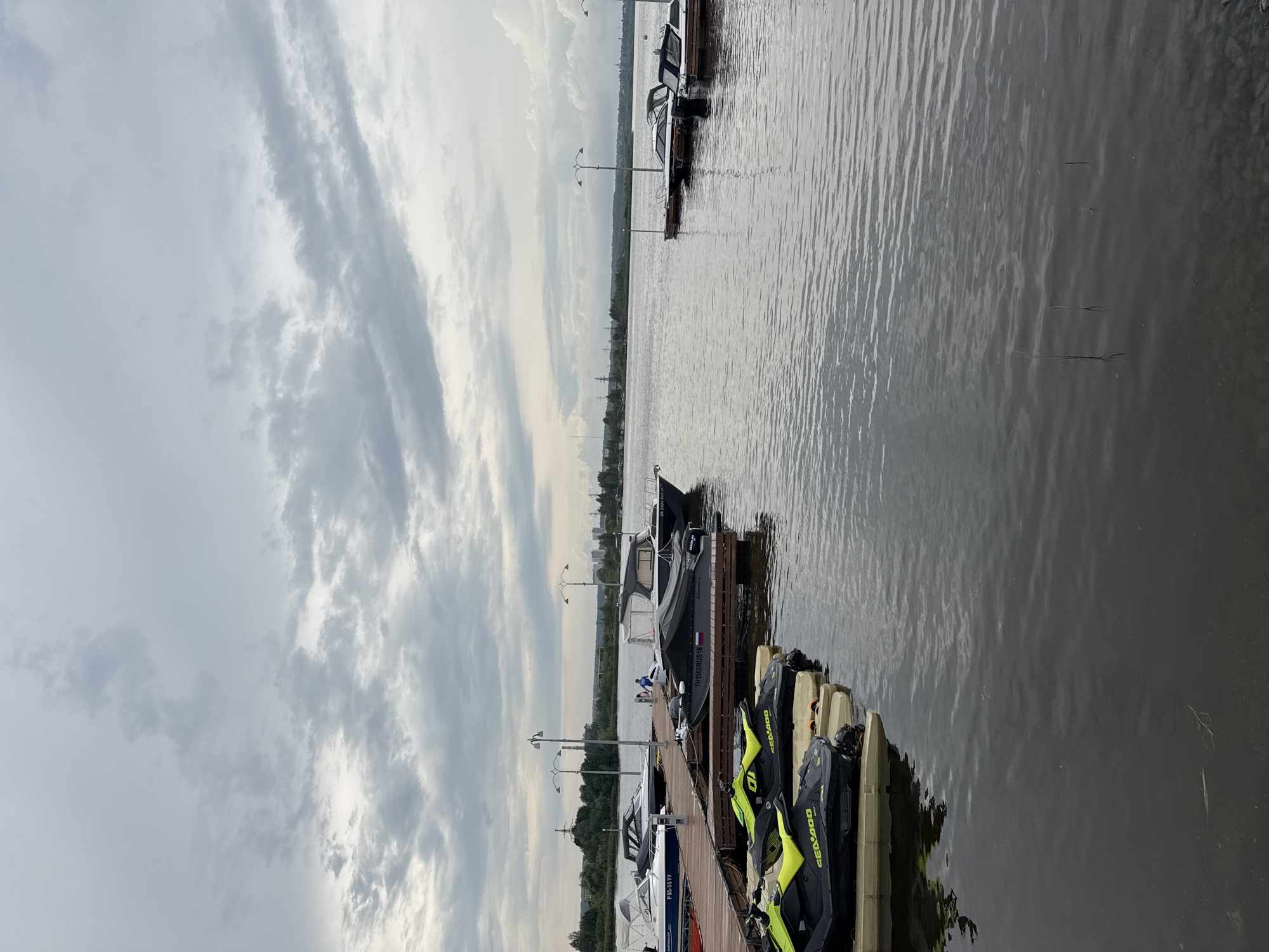 A cloudy day at a marina in Izhevsk, Russia. Several jet skis are docked in the foreground, with their bright yellow seats visible. Behind them, a wooden dock extends into the water, with several motorboats moored alongside. In the distance, the shoreline is lined with trees, and buildings and industrial structures can be seen. The water is calm, with ripples reflecting the overcast sky.