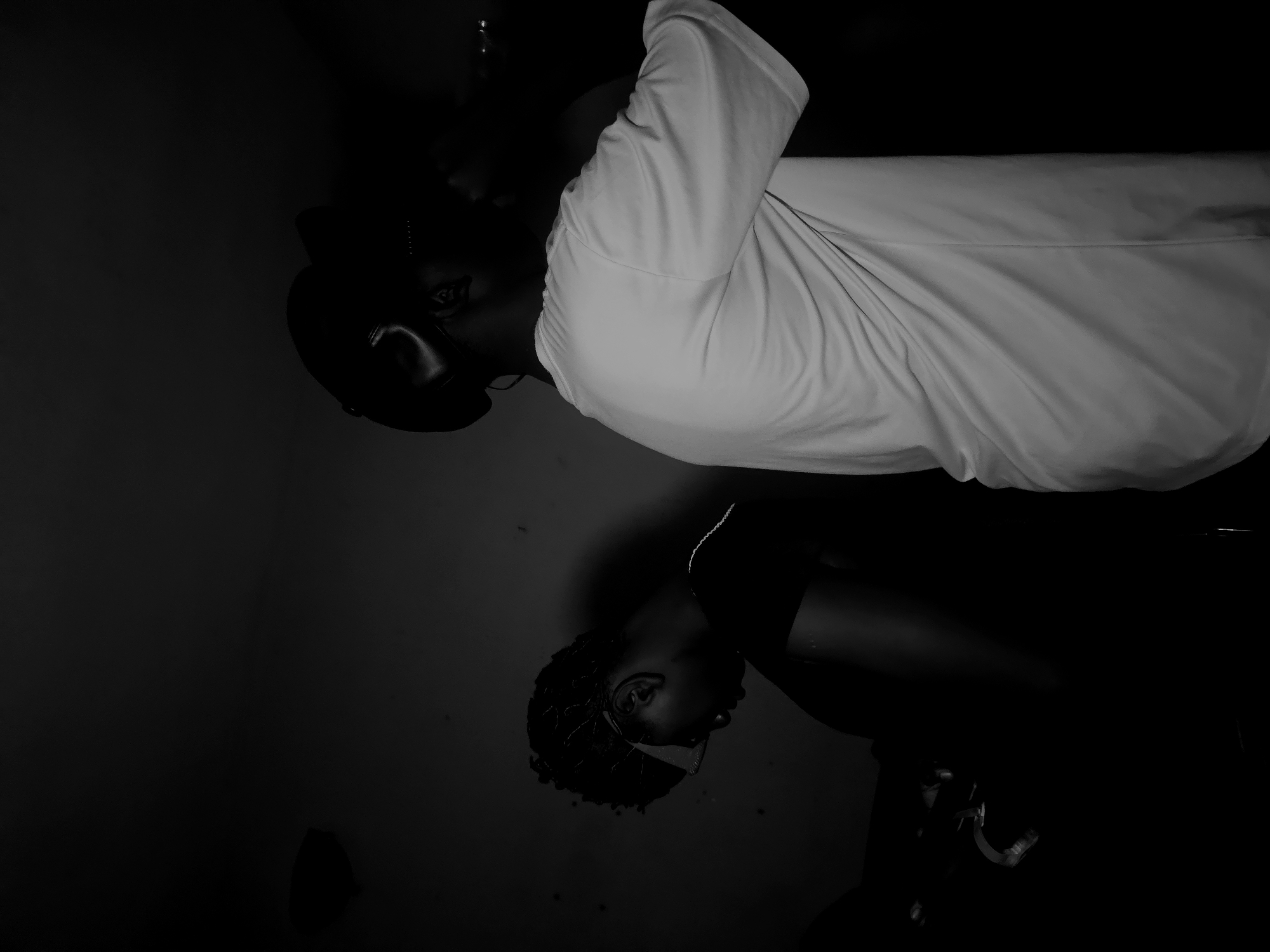The image captures two individuals in a dramatically lit, monochrome setting. The prominent subject, positioned towards the upper right (when the original image is viewed upright), is a person with dark skin wearing a bright, possibly white, short-sleeved shirt. Their head is slightly tilted, and one eye and parts of their face are illuminated, contrasting with their dark hair or head covering and the deep shadows surrounding them.
A second individual is visible in the lower left portion of the image, largely obscured by darkness. This person also appears to have dark skin and textured, possibly curly, hair. They are wearing a dark top, and only an ear and the side of their face are faintly visible due to minimal light. Their head is tilted downwards or away.
The overall scene is characterized by extreme low light, creating strong contrasts and extensive deep shadows that dominate the frame. The background is uniformly black, offering no discernible details about the specific location. The composition features the two figures positioned diagonally, with a large expanse of darkness between them. No specific activity or event is taking place beyond the subjects posing for a portrait. The mood is contemplative or serious, underscored by the dramatic lighting. There is no visible text in the image. The time of day is likely night, or the photograph was taken indoors in a dimly lit space.
