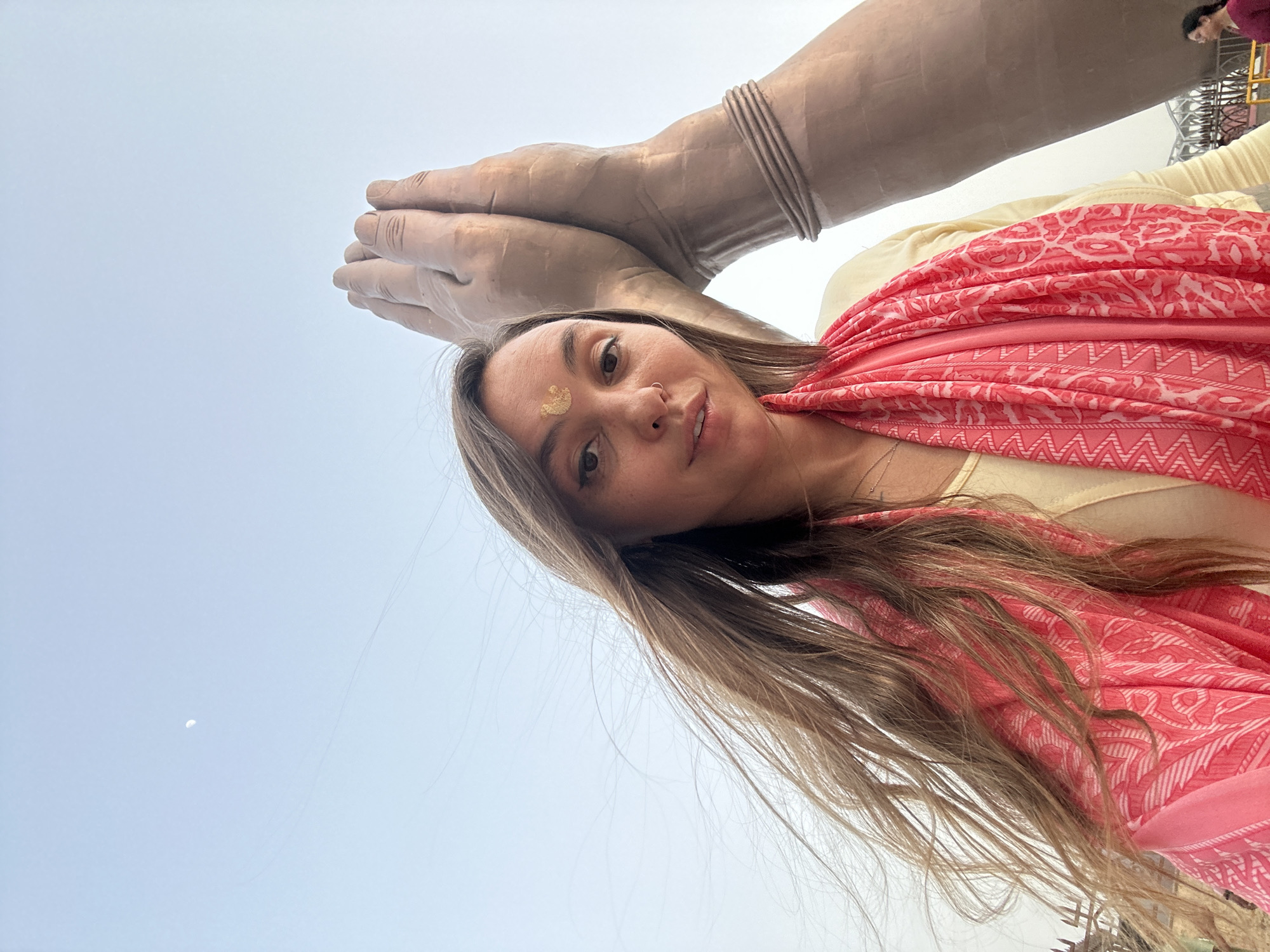 A selfie taken at a low angle features a woman in the foreground, angled towards the upper left of the frame. She has long, light brown hair, a nose ring, and a golden mark on her forehead. She wears a light-colored top and a pink scarf with a patterned design. Above and behind her are two large, light brown hands held in a prayer position, possibly part of a statue. The background is a light blue sky. Parts of a structure are visible near the top right corner. The location is Mughalsarai, India.