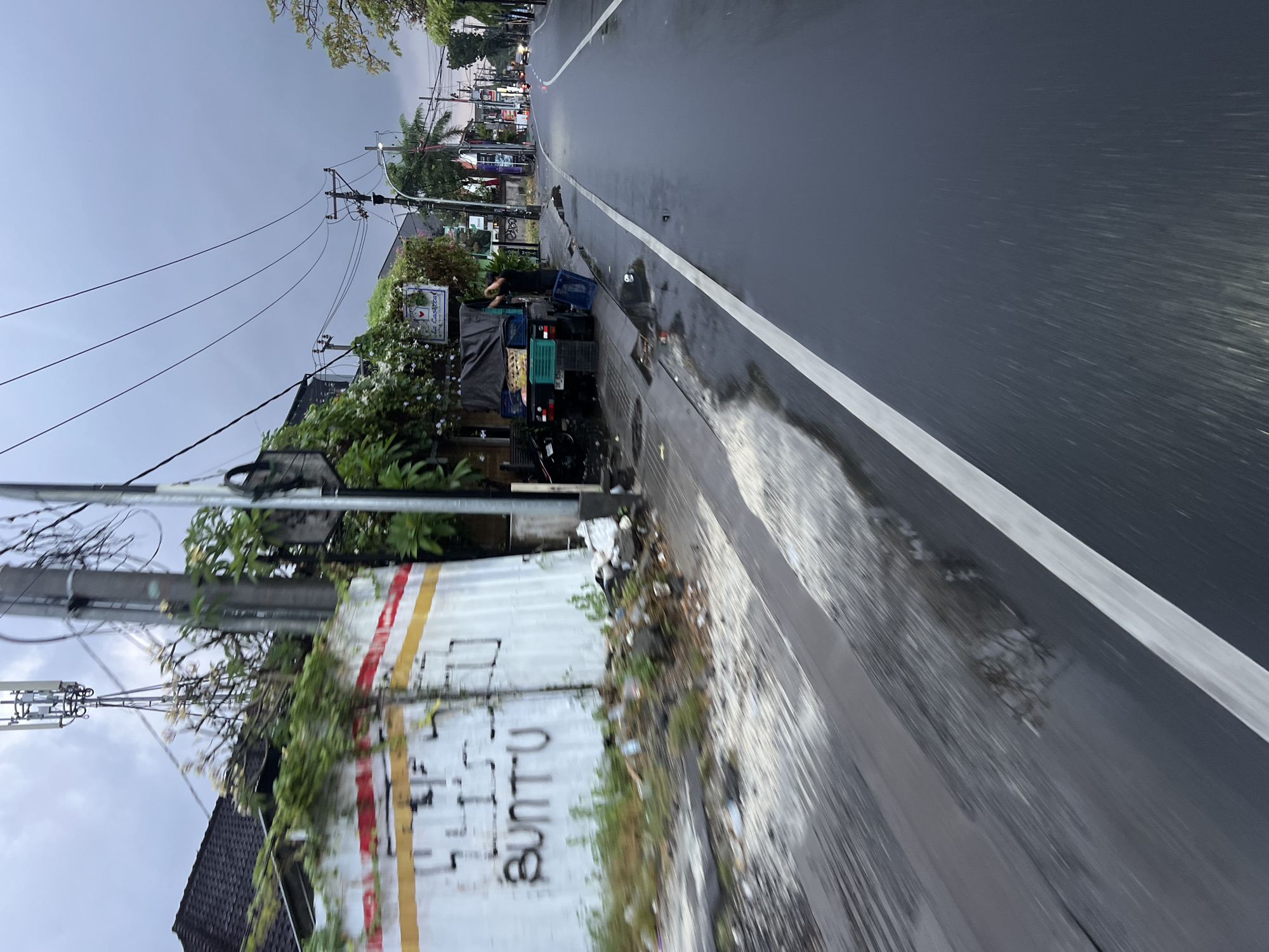 A wet, grey road curves to the right in North Kuta, Indonesia. Power lines crisscross the overcast sky. A white wall, adorned with red and yellow stripes and graffiti reading "BUNTU," lines the left side of the road. Trash is scattered along the base of the wall. A man is visible near a small truck laden with green crates, parked on the sidewalk. Further down the road, a few vehicles are visible in the distance, their lights illuminated. The overall scene suggests a cloudy or rainy day in a somewhat cluttered urban environment.