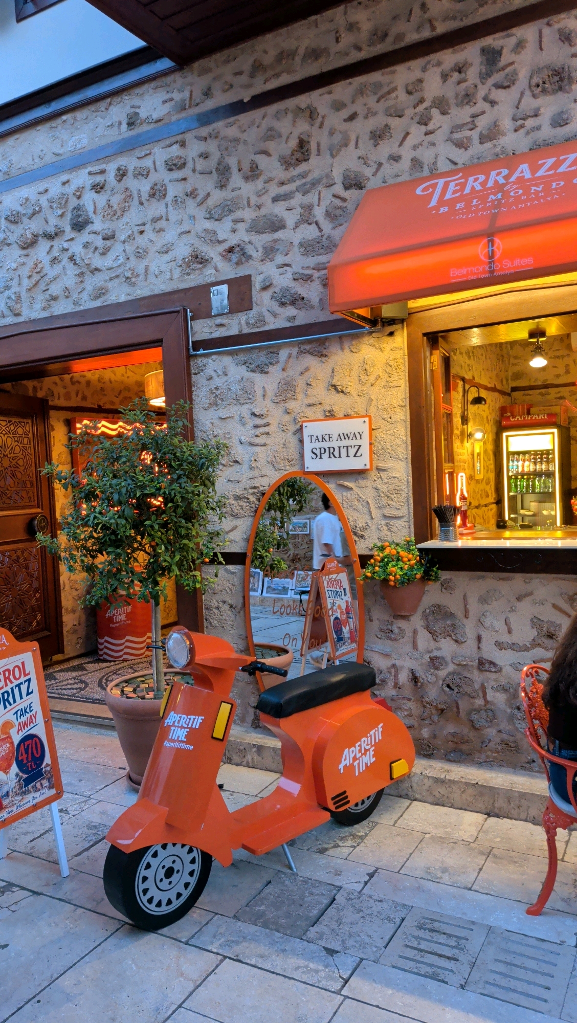 The image captures an outdoor scene in Antalya, Turkey, likely in the Old Town (Kaleici) area, given the traditional stone architecture. In the foreground, a bright orange scooter, decorated with "APERITIF TIME" branding, is parked on a cobblestone street. To the left of the scooter, a sign advertises "APEROL SPRITZ" with a price of "470 TL". A potted tree with lush green leaves stands to the left, and a large, ornate oval mirror leans against the stone wall.
Above the mirror, a sign reads "TAKE AWAY SPRITZ". The building facade is constructed of rough-hewn stones, with wooden doorways and window frames. An orange awning extends from the building above a service window, displaying the text "TERRAZZO BELMONDO SUITES OLD TOWN ANTALYA". Inside the service window, a bartender is visible, and a refrigerated display case filled with bottles of Campari is illuminated. Orange flowers in a pot are placed on the counter. To the far right, a person is seated at a red wrought-iron chair, facing away from the camera. The overall atmosphere suggests a vibrant and inviting establishment.