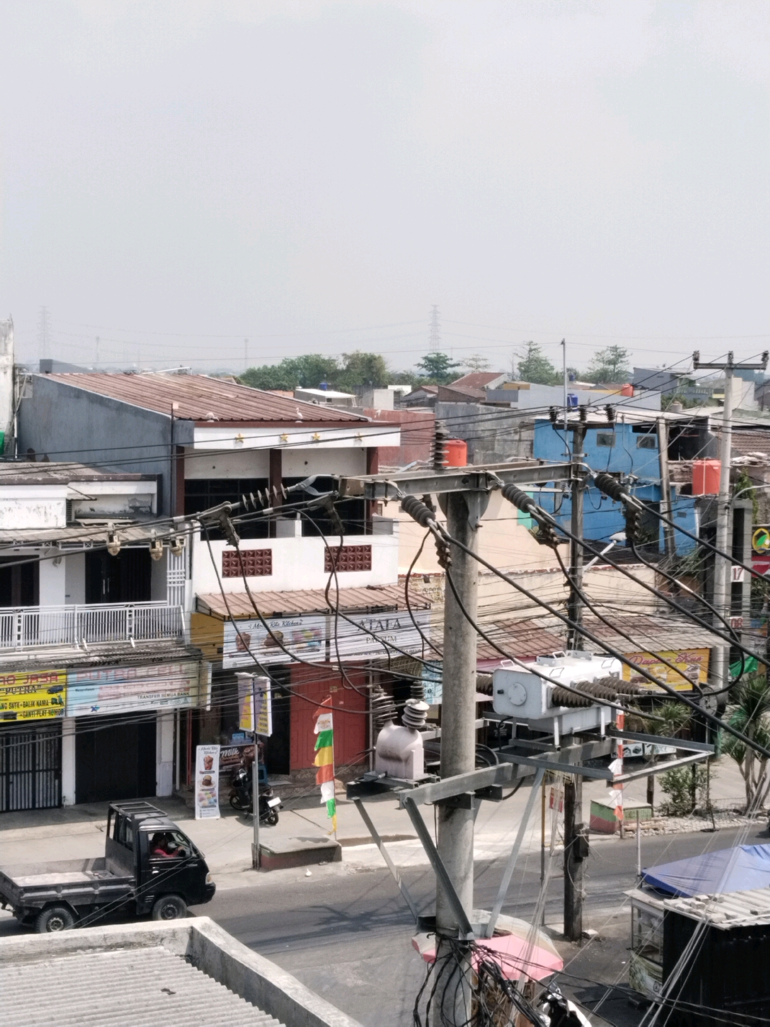 The image depicts a street scene in Jalenjaya, Indonesia, under a hazy sky. In the foreground, a black pickup truck with a driver is on the road. To the left, a building with a white facade and a balcony is visible, with signage for "BUANA JASA". Adjacent to it is another building with a dark facade and various advertisements. Further down the street, a white utility pole with numerous electrical wires, transformers, and insulators dominates the mid-ground. Behind the utility pole, a row of shops and houses with corrugated metal roofs can be seen. A bright blue building with red water tanks on its roof is prominent amongst them. To the right of the utility pole, a smaller red and green structure is present, and further to the right, a blue tarp covers a stall. Trees are scattered throughout the background, and a distant electrical tower is visible on the horizon. The overall impression is of a busy, densely populated urban or suburban area with ongoing electrical infrastructure.