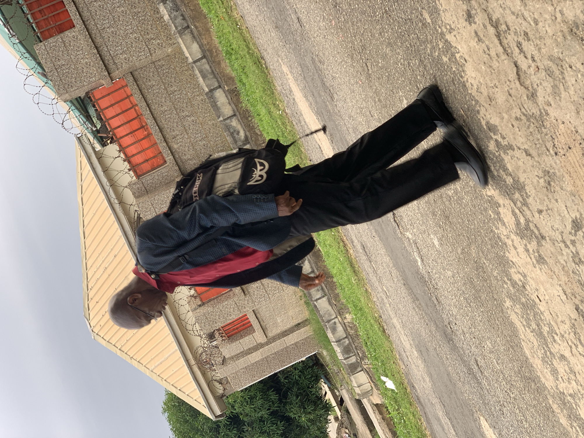 In an outdoor scene in Abuja, Nigeria, a Black man is walking on a paved road, viewed from the side and slightly from above. He is wearing a red shirt, a blue patterned jacket, and black trousers. A backpack with a logo is slung over his right shoulder. The man is wearing glasses and appears to be bald. In the background, a building with a yellow roof and a stone facade is visible. The building is surrounded by a fence topped with barbed wire and has red-colored gates or window coverings. Green grass lines the side of the road. The sky is overcast.