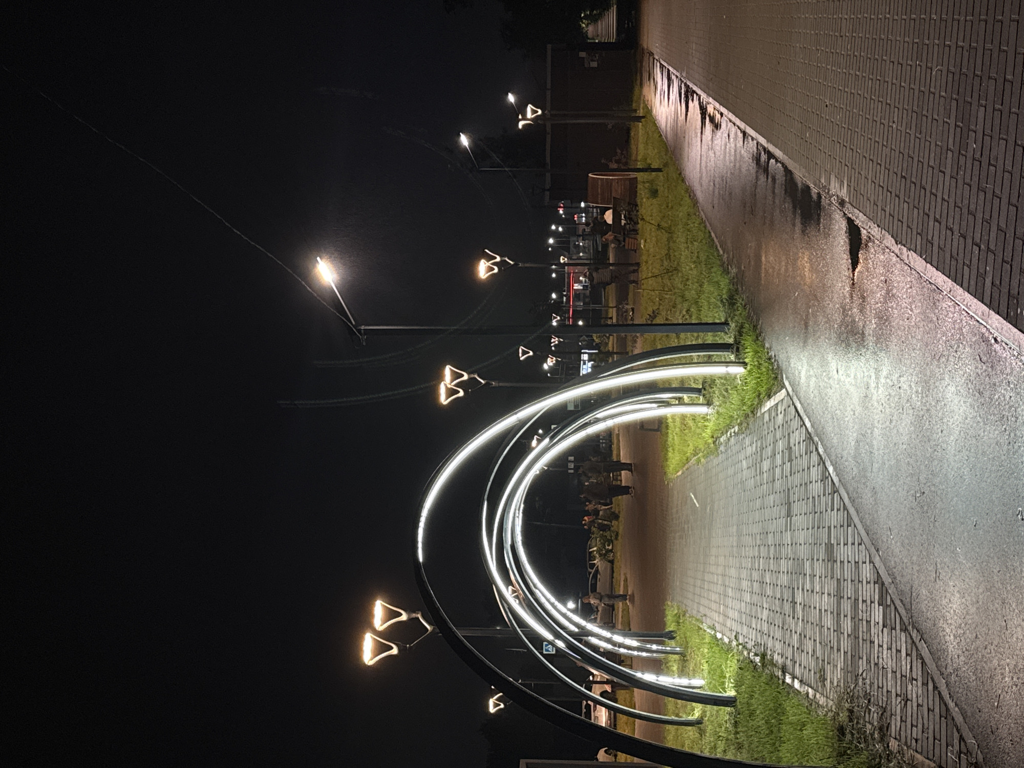 The image depicts a nighttime outdoor scene in Chaikovsky, Russia, characterized by artificial illumination after recent rain. A prominent feature is a series of five successive black metal arches, internally lined with white LED lights, spanning a grey paver pedestrian walkway that recedes into the background. The walkway surface appears wet and subtly reflective.
To the right of the arched pathway, a parallel, wider paved area consists of dark brick pavers adjacent to a wet, reflective asphalt or concrete section with visible puddles. This path also extends into the background.
Numerous streetlights illuminate the scene. Several lights feature distinctive dual, upward-facing, "W"-shaped fixtures emitting warm orange-yellow light. Other streetlights, particularly on the far right and mid-left, have single, conventional fixtures emitting cooler white light. Faint, thin lines, possibly power lines or wires, are discernible against the dark sky.
Approximately 8-10 individuals are present in the mid-ground. Several people are standing or walking near the end of the arched walkway. On the right, a group gathers near a cylindrical wooden structure, possibly a bench, with at least two individuals seated. In the background, through the arches, multiple parked vehicles are visible, including a white car on the left. Distant lights suggest commercial establishments. Grass verges separate the pathways and are visible along the sides of the arched walkway. A dark, possibly brick building or structure is partially visible on the far right.