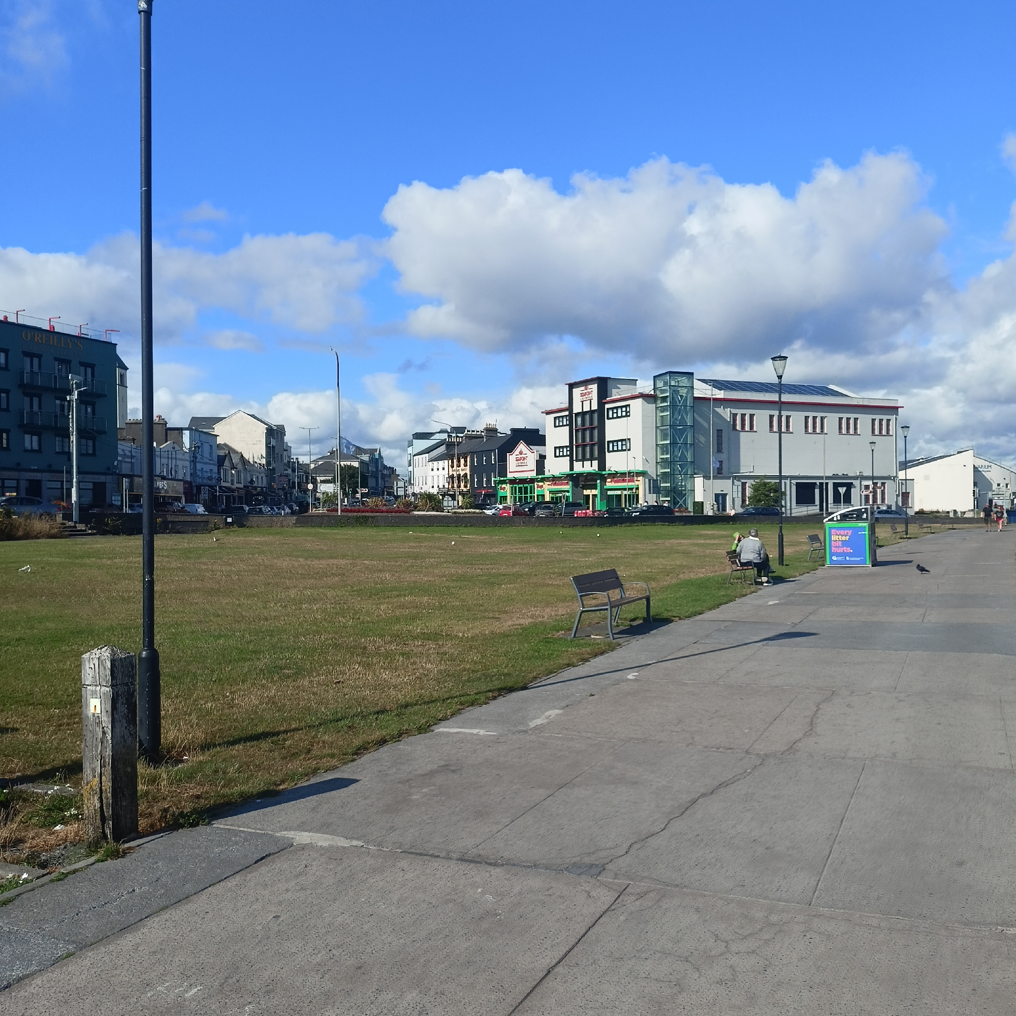 This is a photograph taken on a sunny day with blue skies and fluffy white clouds, likely in the city of Galway, Ireland. The image shows a promenade or walkway in the foreground, paved with concrete slabs. To the left of the promenade is a grassy area, likely a park or public green space.
Along the promenade, there are several benches, one of which is occupied by a person sitting down. Further down the promenade, a green and blue public litter bin with the message "Every litter bit hurts" is visible. A pigeon is standing on the pavement near the bin.
In the background, a row of buildings lines the street beyond the grassy area. These buildings appear to be a mix of businesses and residences, with various architectural styles. One prominent building is a modern hotel or commercial complex with a glass elevator shaft. There's also a petrol station with green and white branding visible. A tall black lamppost stands on the promenade, and several other lampposts are visible along the street. A wooden post with a yellow marker is positioned at the edge of the grass. The overall impression is of a well-maintained urban area, possibly near the coast, given the clear sky and open space.