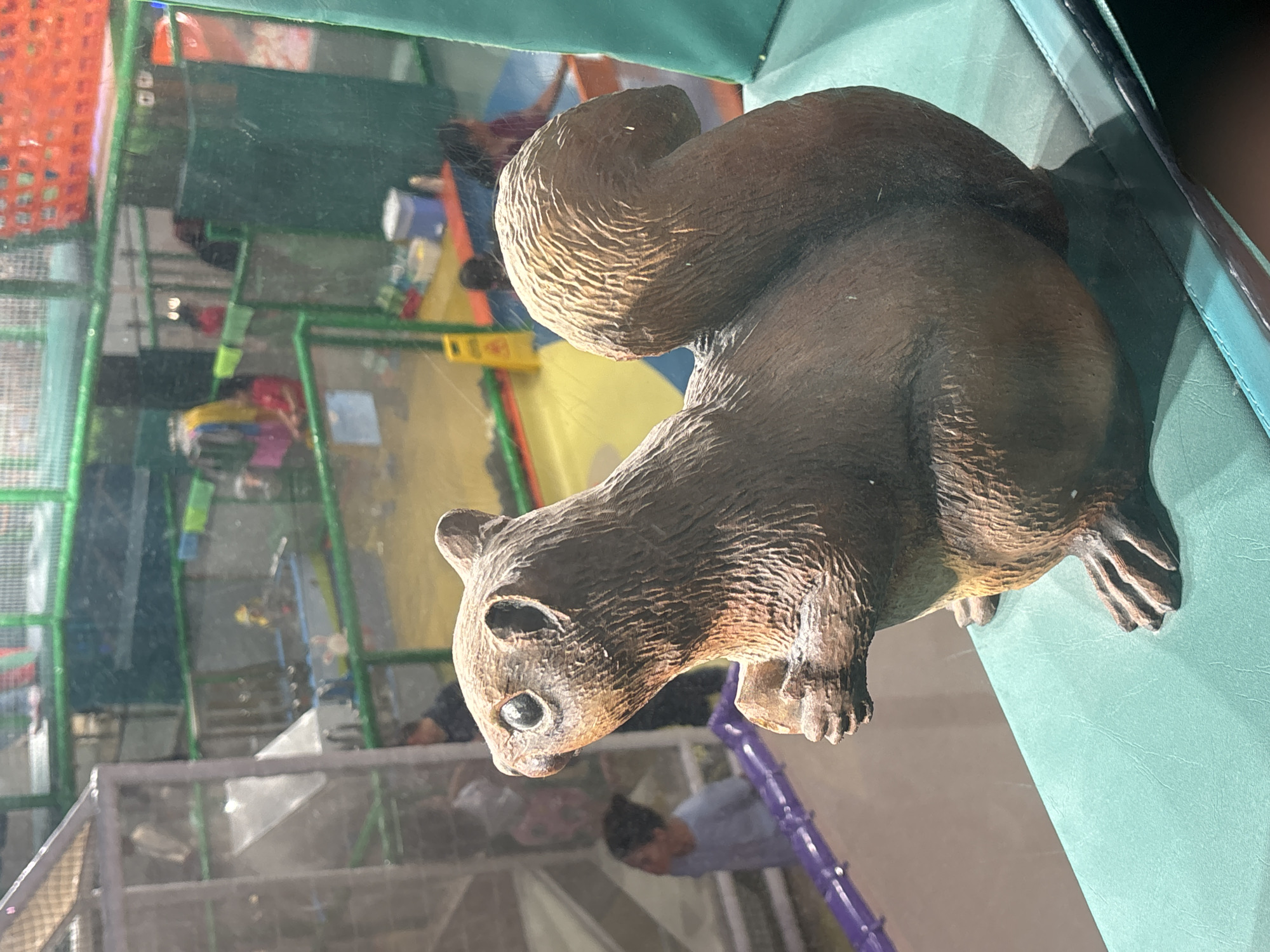 An artificial brown squirrel statue, resembling a carving, is positioned on a teal-colored padded surface in the foreground. The squirrel is depicted holding a nut in its paws and its tail curls upwards. In the background, through a transparent barrier that appears to be glass, a children's indoor playground is visible. This playground features colorful structures with green netting and multiple levels. Several children and adults are present in the background, engaged in various activities within the play area. The overall scene suggests a lively environment designed for recreation. The location is an indoor children's play area, likely within Dubai, United Arab Emirates.