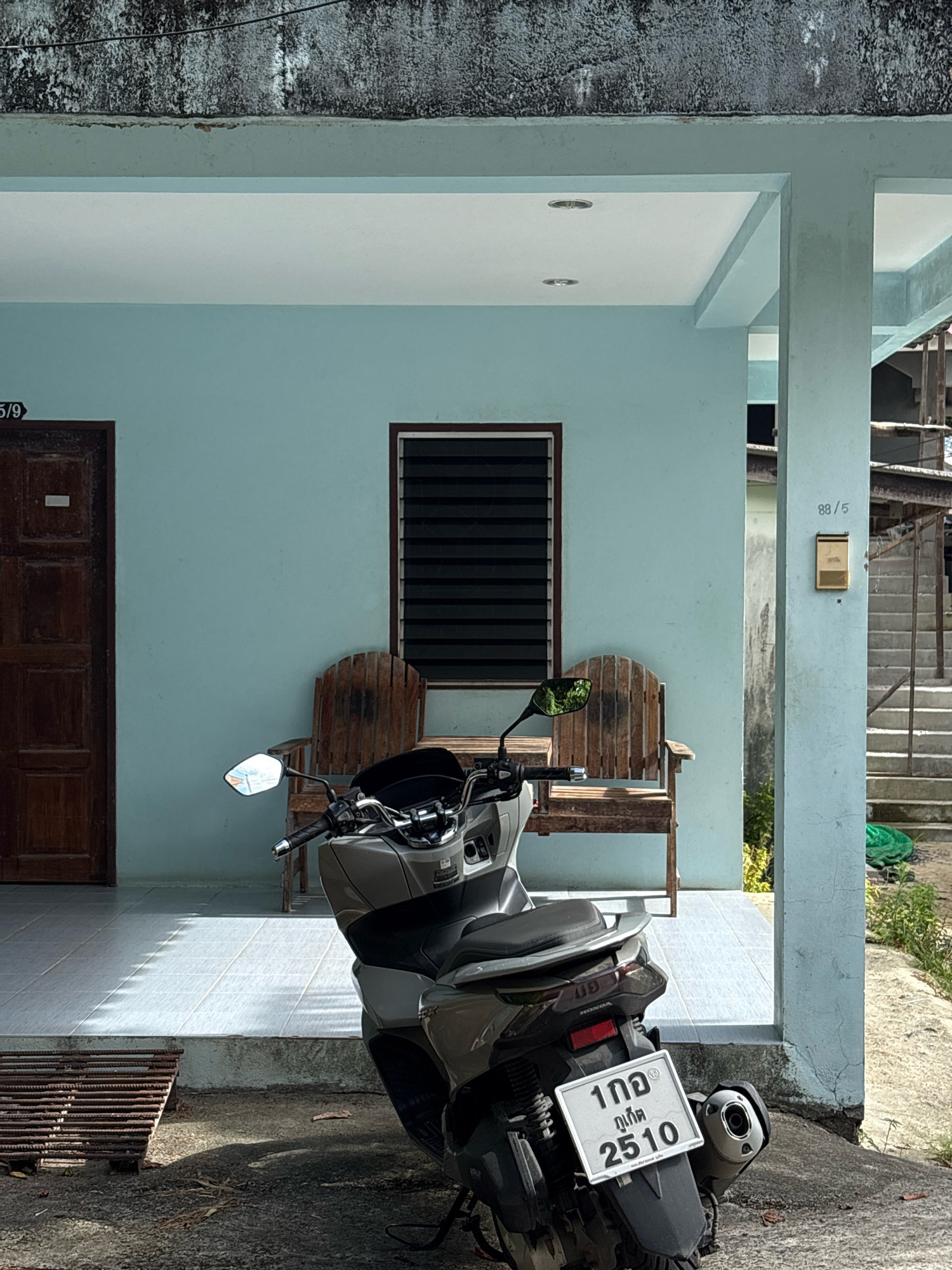 The image depicts a scooter parked in front of a light blue building with a covered porch area. The building appears to be an older structure with some wear and tear on the exterior, particularly the concrete ceiling. To the left, a dark brown wooden door is visible with a small white sign above it. To the right of the door, a window with horizontal blinds is set into the wall. Two wooden chairs with slatted seats and backs are placed on the tiled porch.
The scooter is a modern-looking, grey-colored model, positioned centrally in the foreground. It has a prominent license plate with Thai characters and numbers "1กอ ภูเก็ต 2510". The scooter's exhaust pipe is visible on the right side.
Beyond the immediate building, a concrete staircase leads upwards on the right side, with a metal railing. A golden mailbox is affixed to a support pillar near the stairs, displaying "88/5". The environment suggests a somewhat rural or less urbanized setting, possibly in a residential area. The lighting indicates it is daytime, with shadows cast on the ground. There are no people visible in the image. The overall impression is one of a quiet, possibly residential or small commercial, establishment in a tropical climate, consistent with locations like Choeng Thale, Thailand.