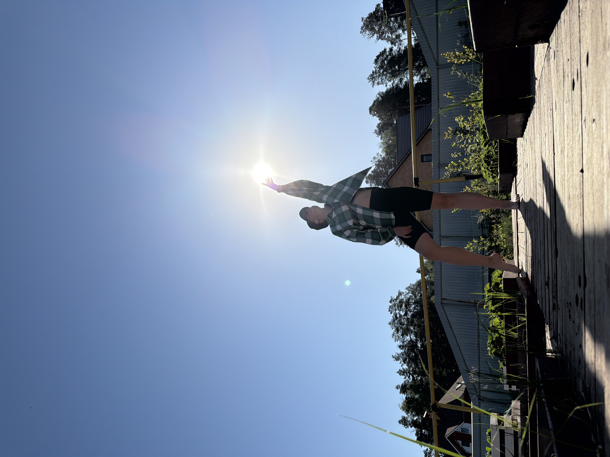 A person stands barefoot on a wooden boardwalk, reaching towards the sun. The person is wearing black shorts and a green and white plaid shirt, open at the front. The boardwalk is adjacent to vegetation and a fence. In the background, a building and trees are visible. The sky is blue. The image was taken during daylight hours and it is likely sunny. The location is in Chaikovsky, Russia.